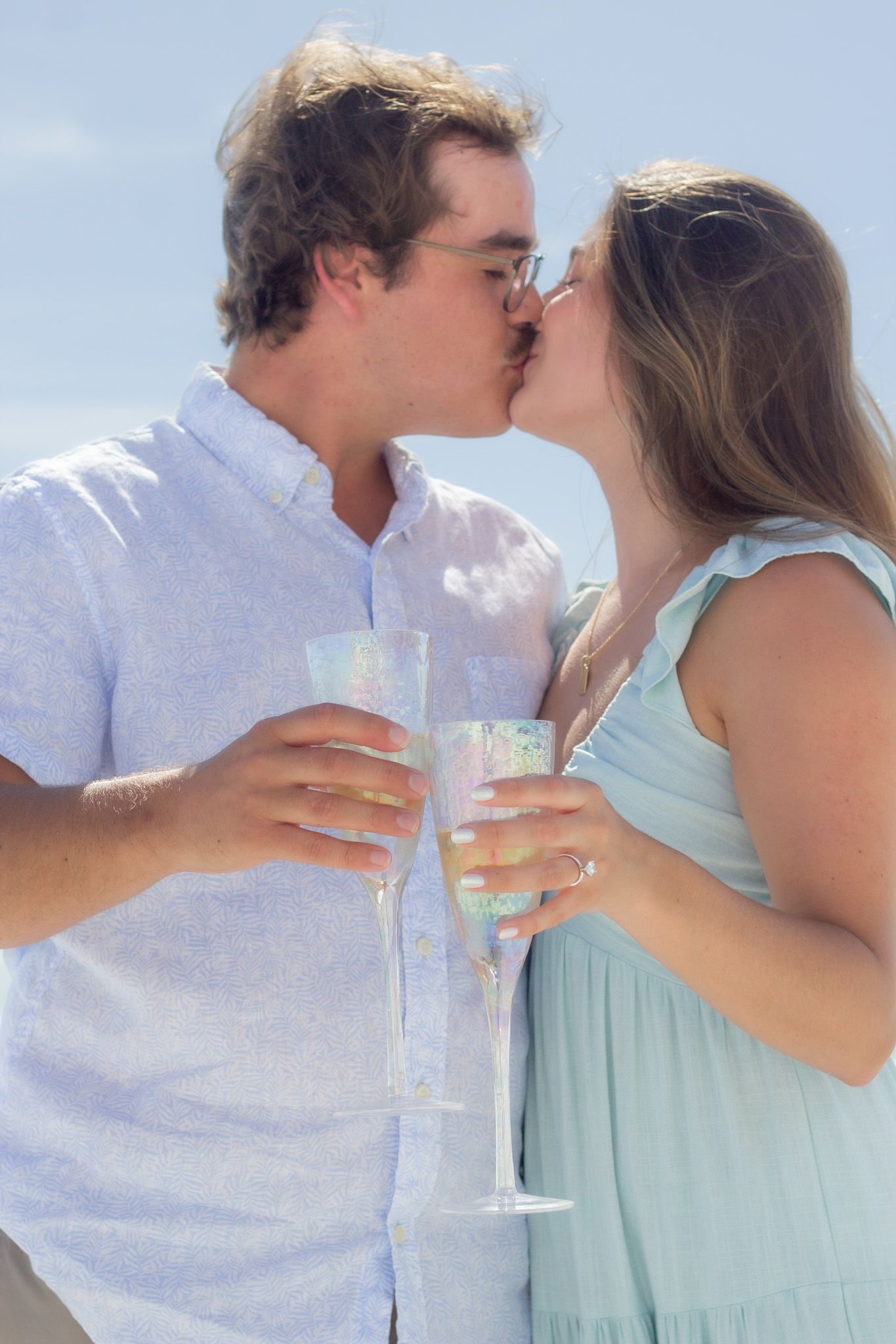 Couple in light summer attire shares champagne toast and kiss during beachside celebration.