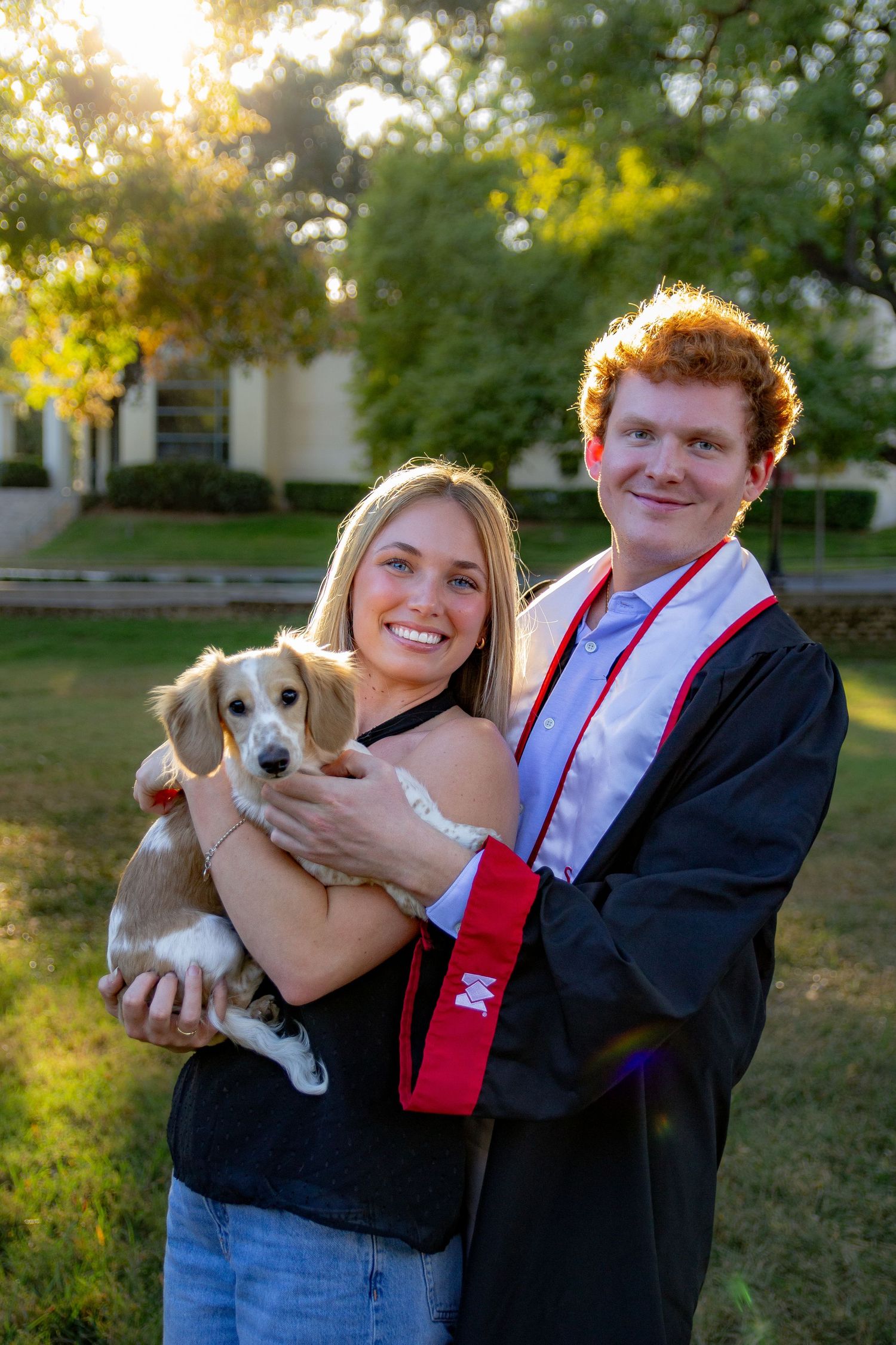 A couple in formal attire holds a small beagle puppy outdoors during golden hour in a park setting.