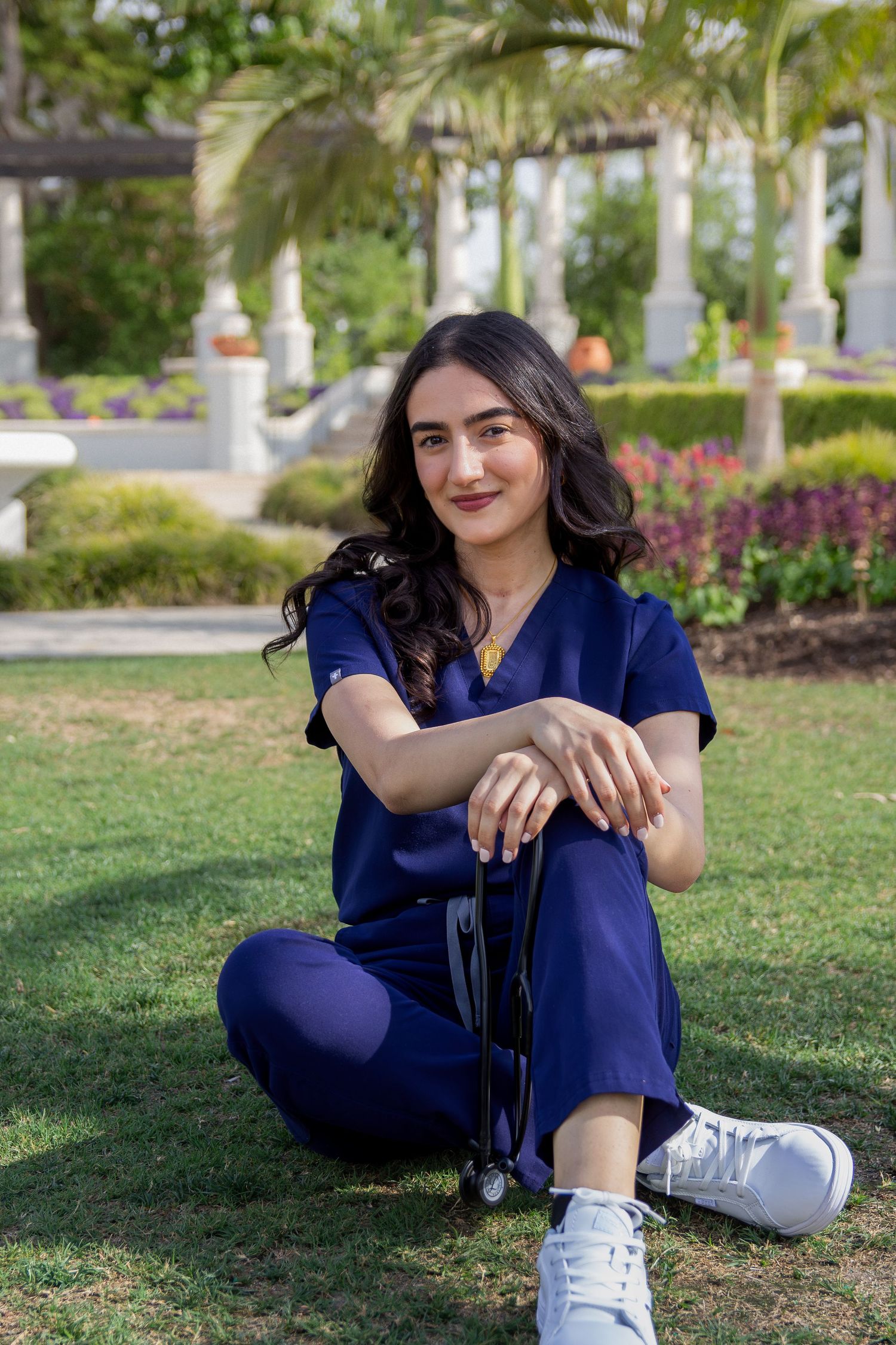Young woman in navy blue scrubs sitting on grass in a garden, smiling confidently with a stethoscope nearby.