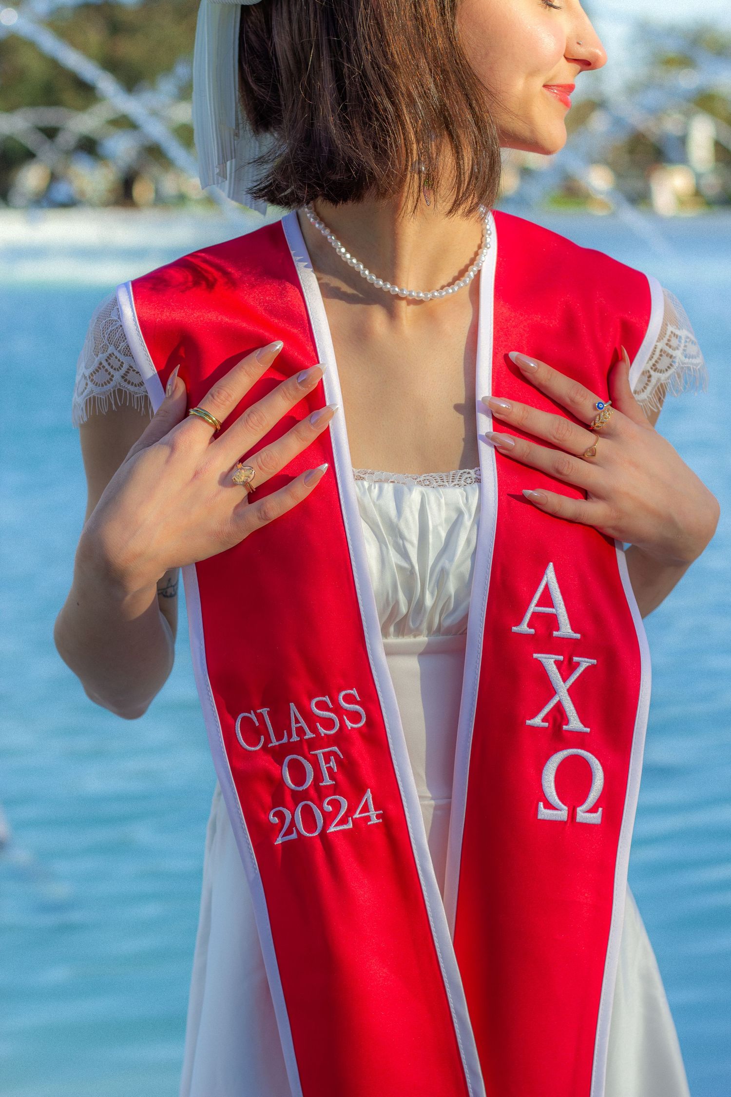 Graduate wearing a red Alpha Chi Omega sorority stole with Class of 2024 lettering poses against blue water background.