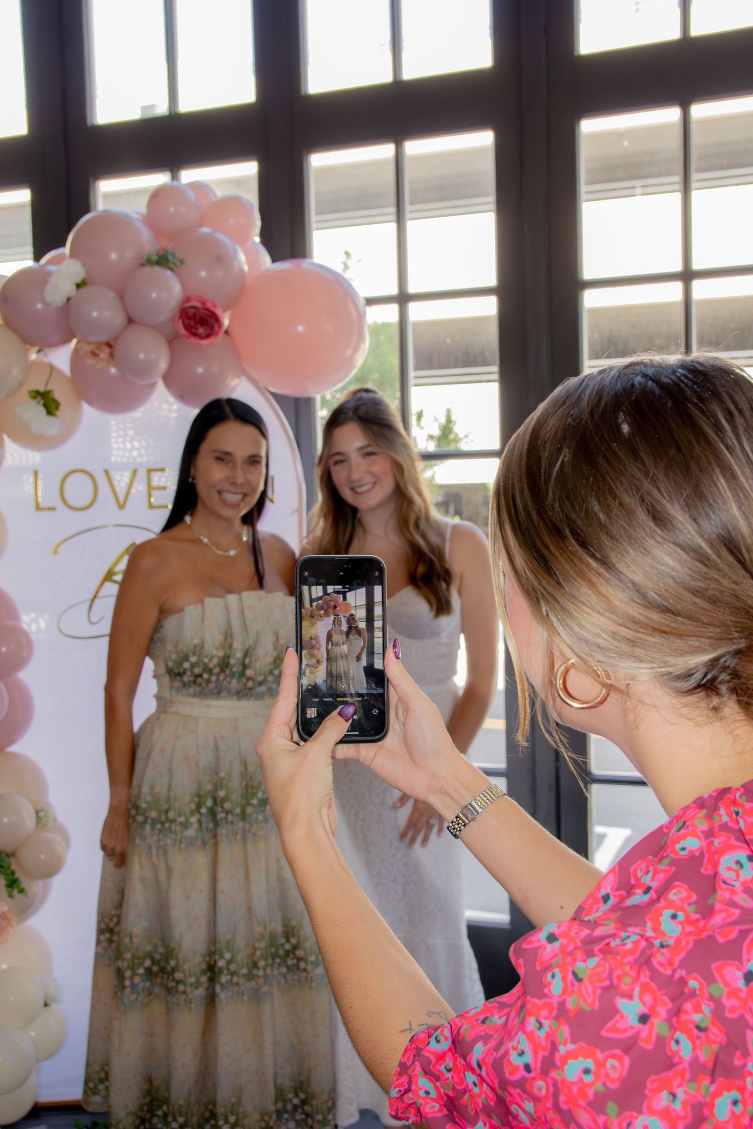 Woman in pink floral top takes photo of two people posing in beige dresses at an event with balloon decorations.