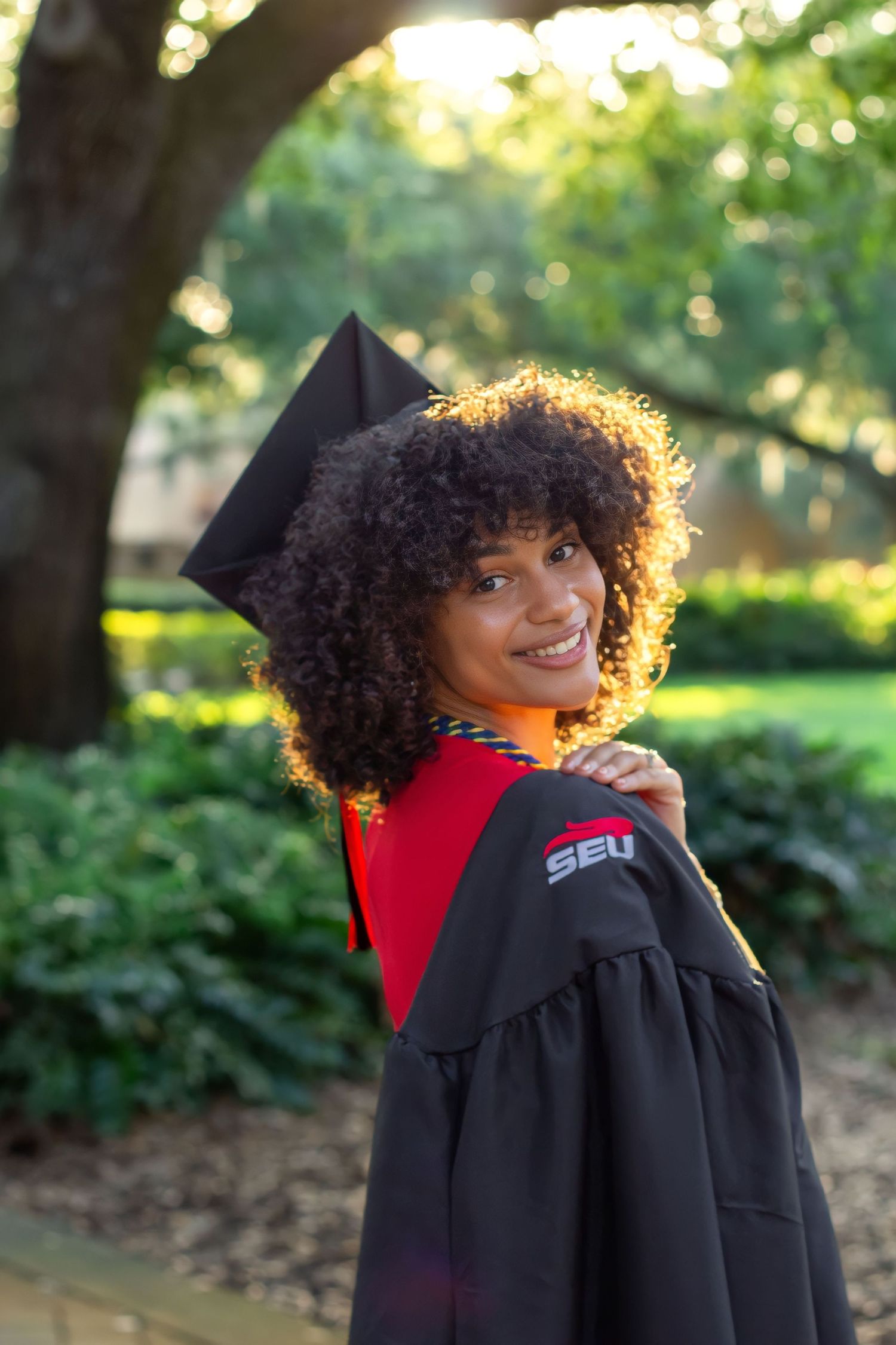 Graduate in black academic regalia with curly hair smiles while looking back over their shoulder in an outdoor setting.