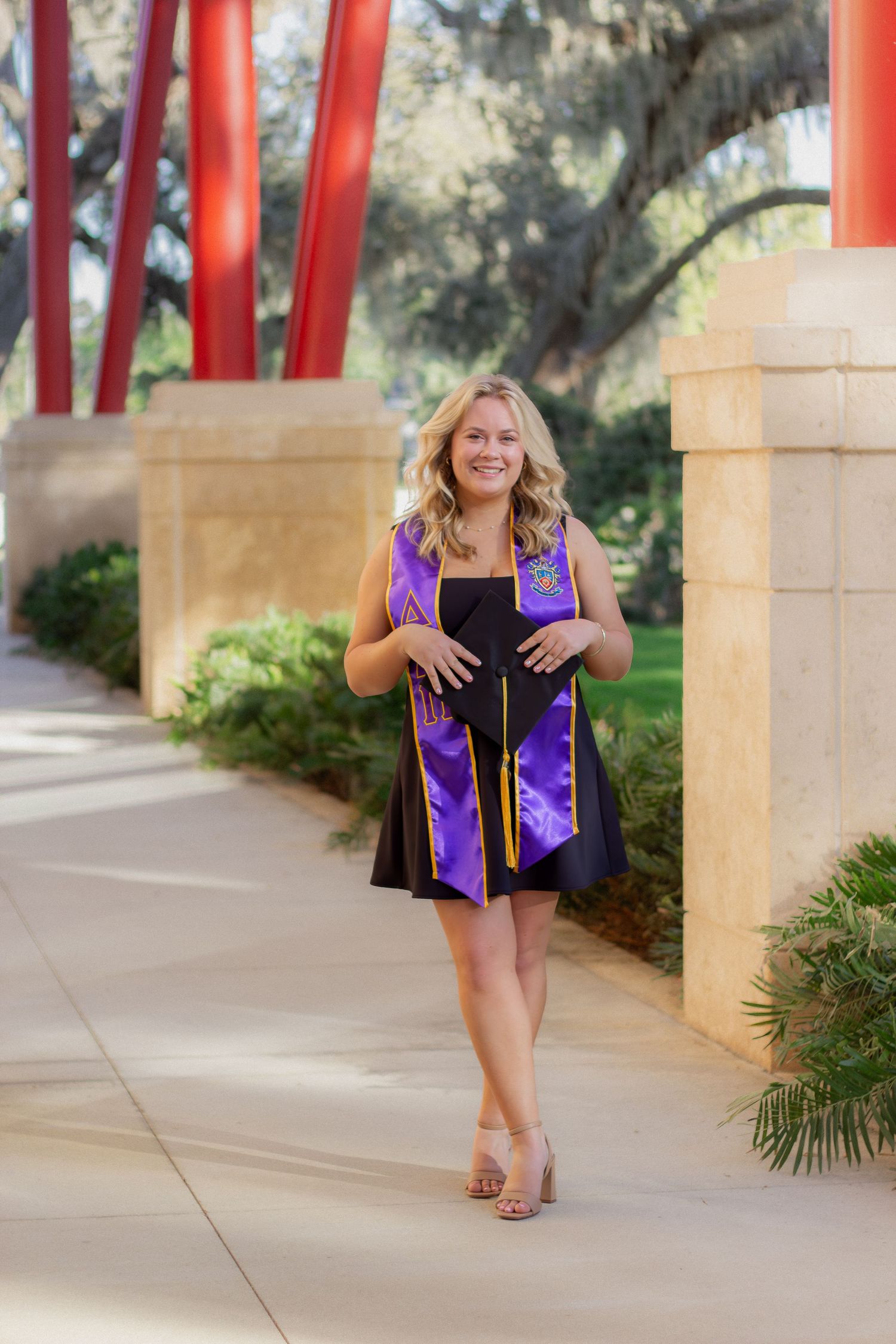 Graduate in a purple and black graduation gown stands on campus walkway with stone pillars and trees.