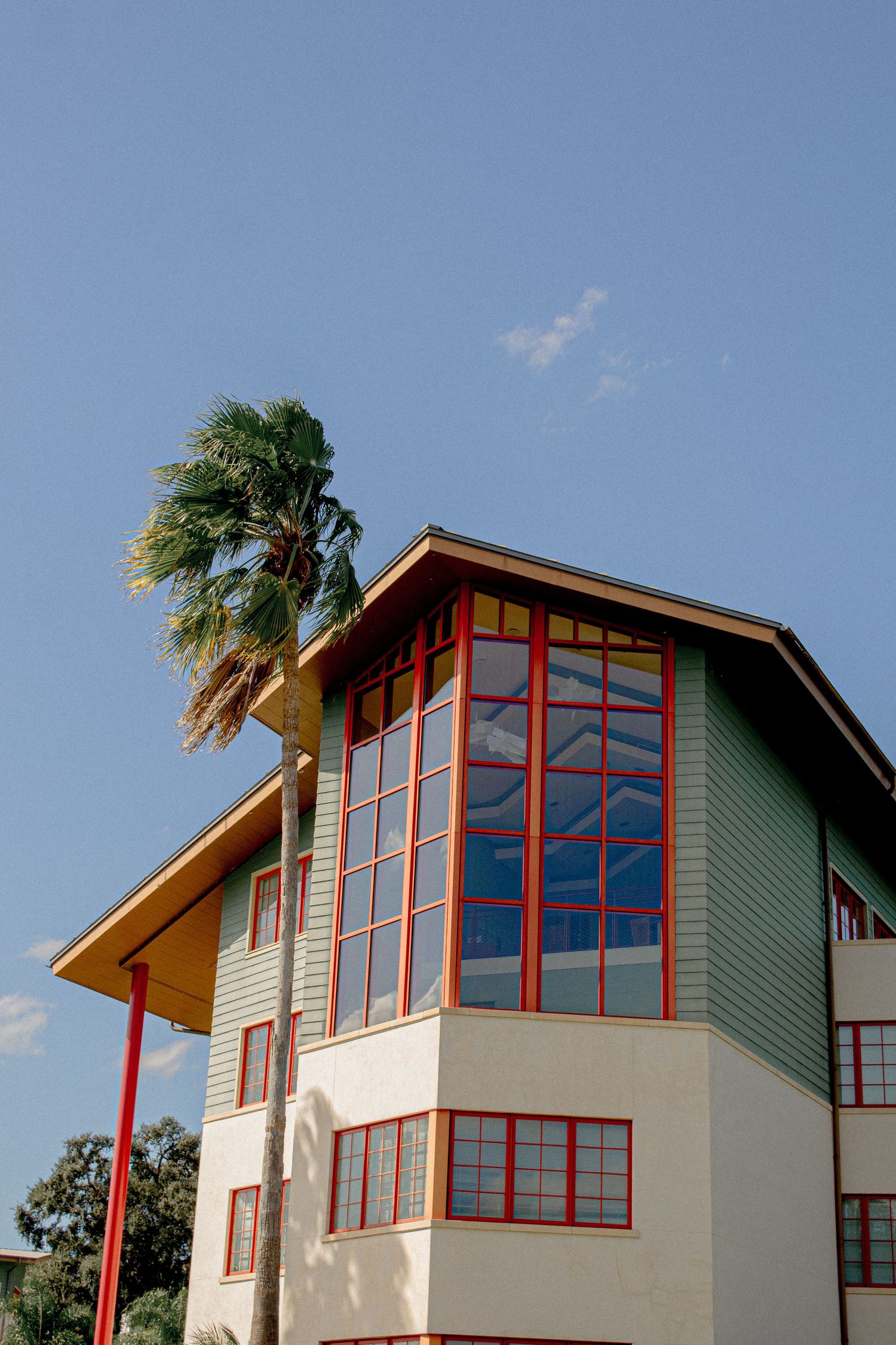 Modern green and white building with large angular windows and red trim features palm trees against a blue sky.