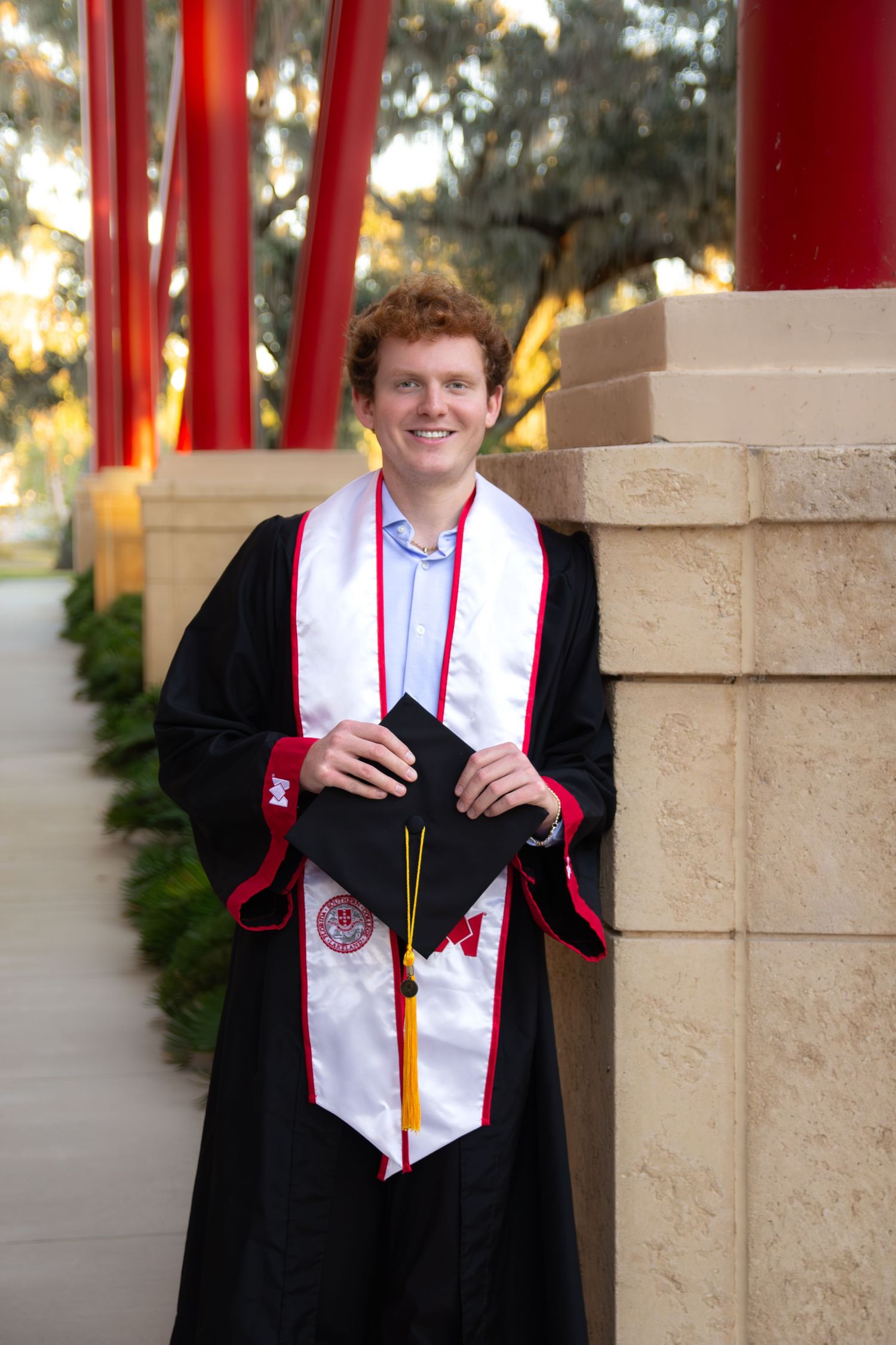 Graduate wearing black cap and gown with red and white stole poses in front of red pillars at sunset.