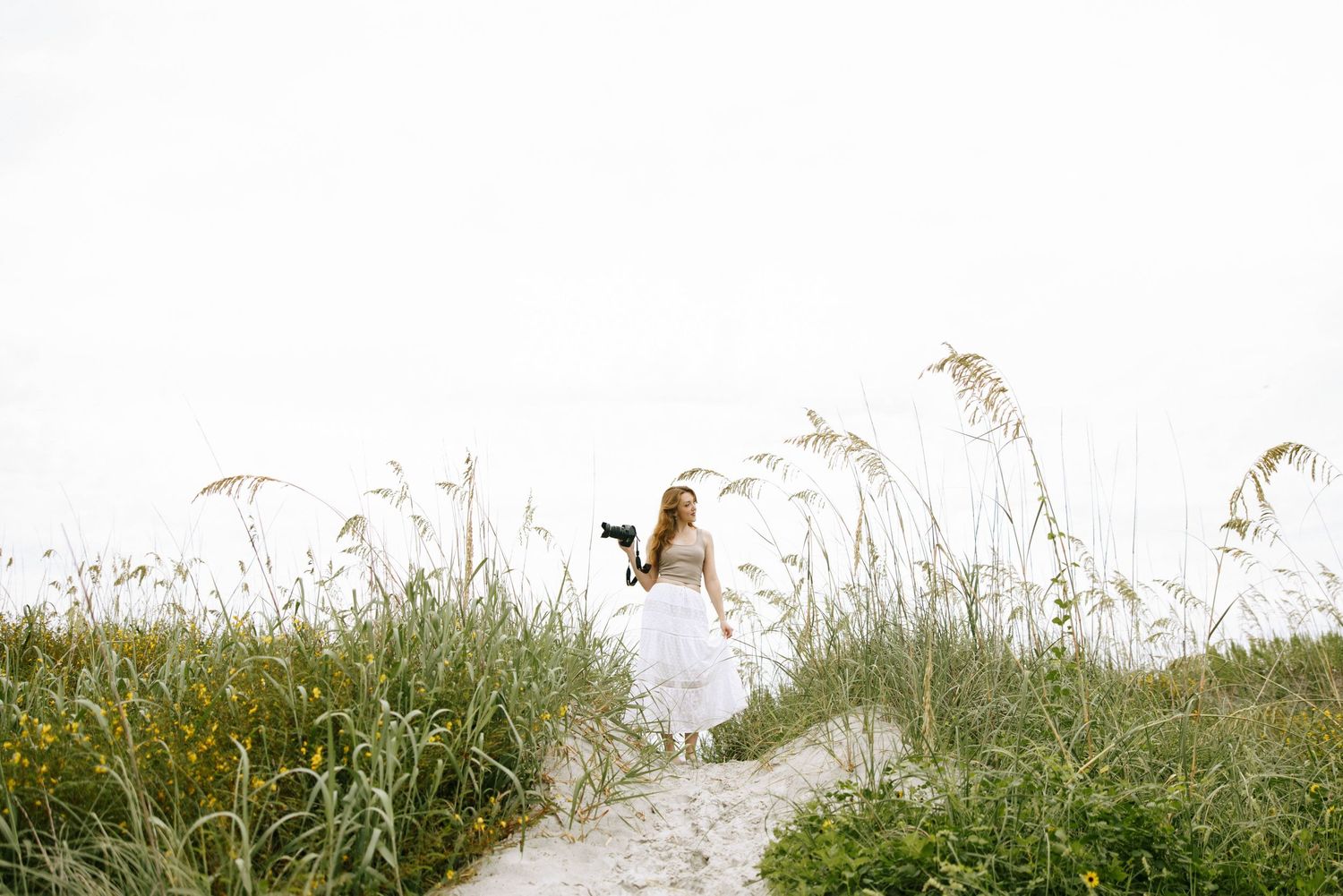 Bride in white gown walking sandy beach path surrounded by tall sea oats grass on a cloudy day.