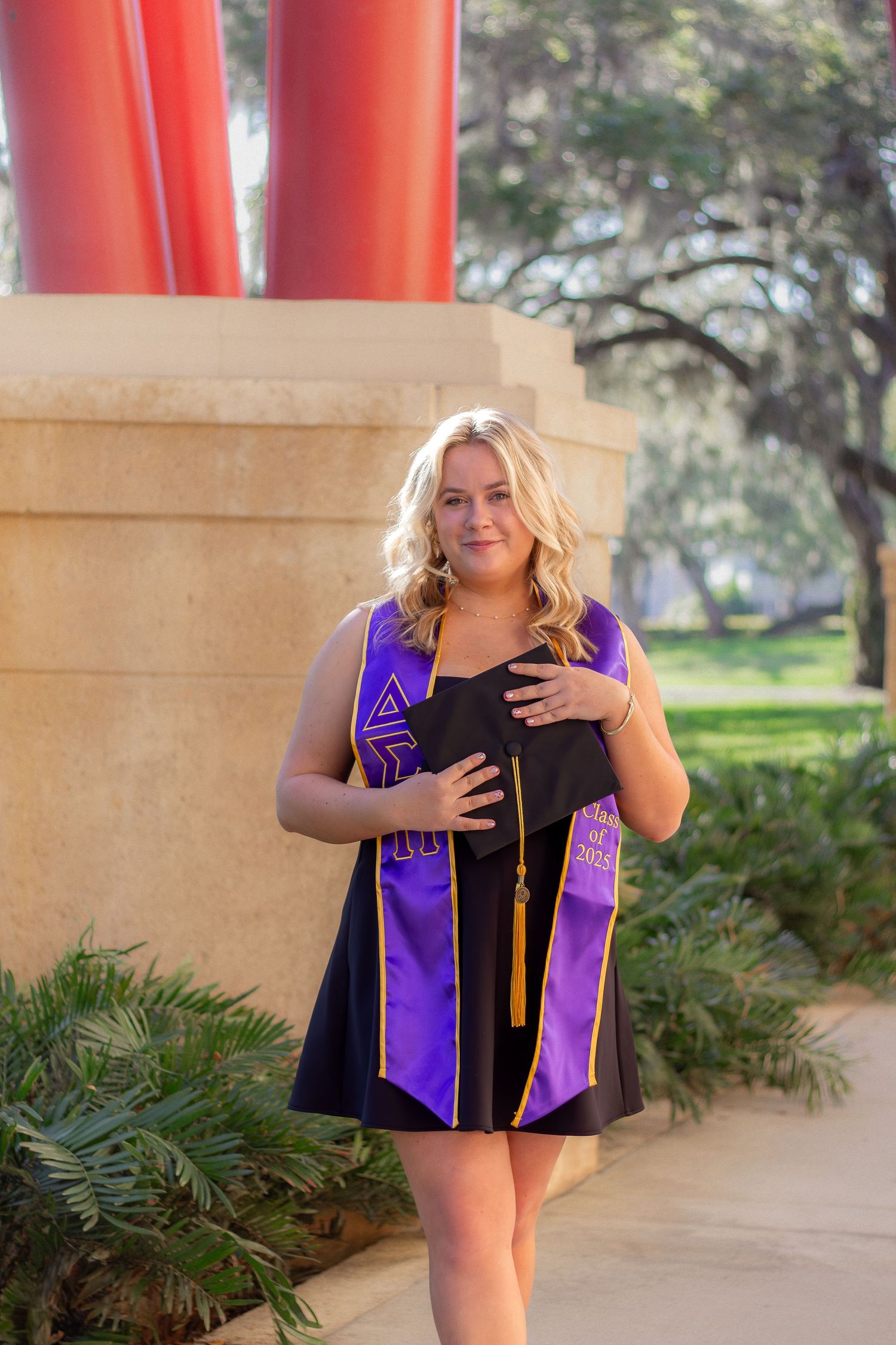 Graduate wearing purple graduation stole and black cap poses in outdoor walkway with columns and plants.