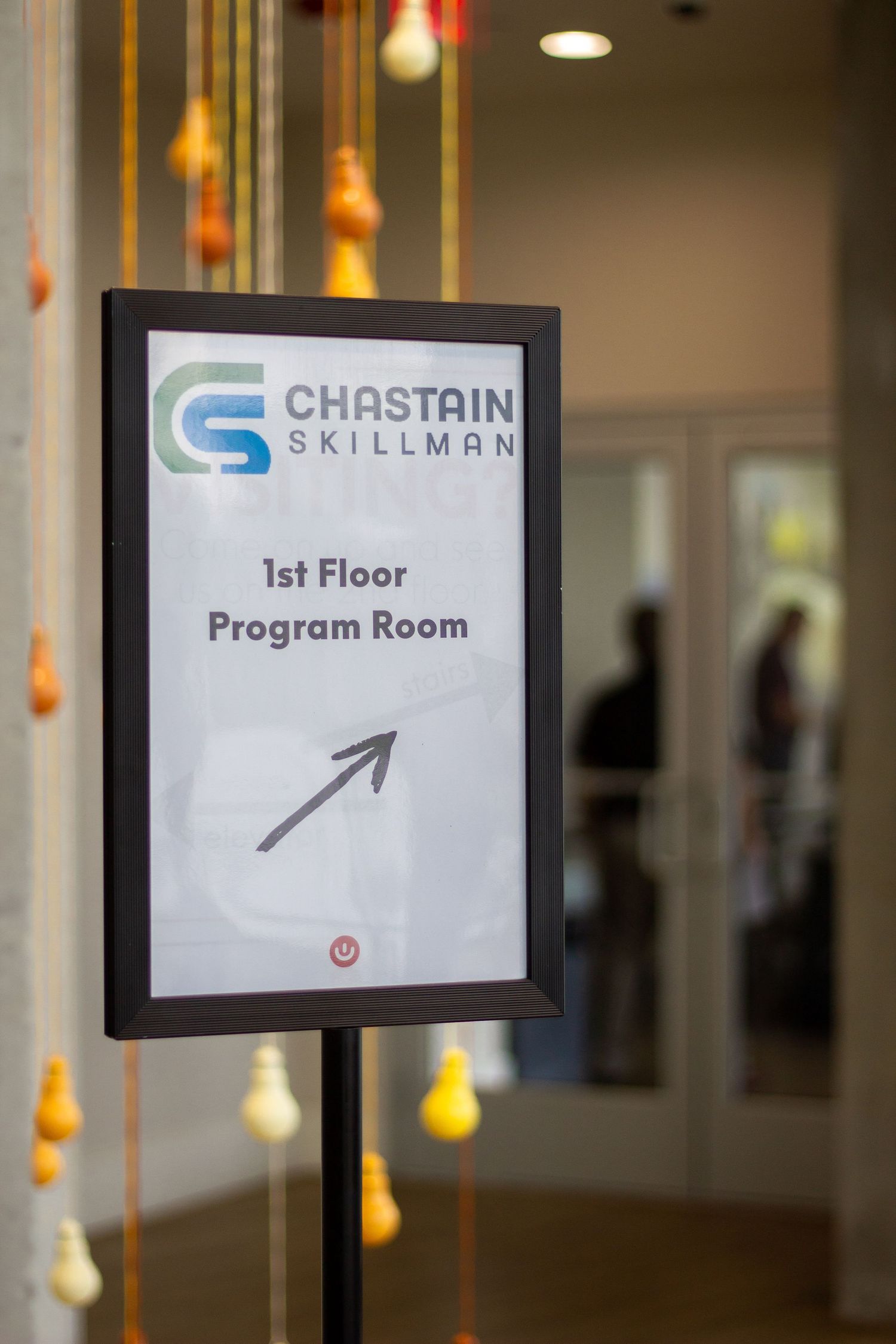 Directional sign for Chastain Skillman's first floor program room with decorative hanging light bulbs in foreground.