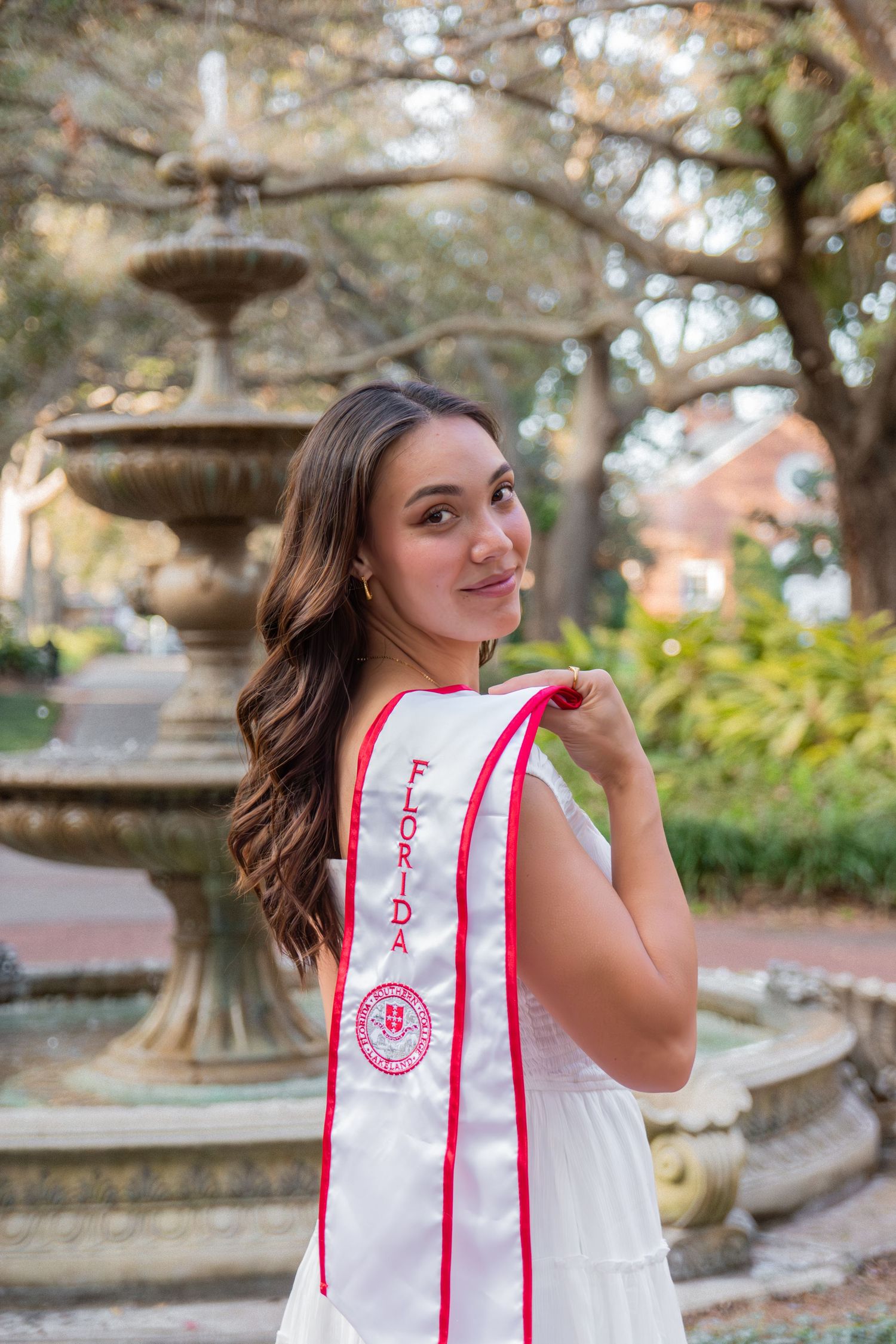 Outdoor graduation portraits near ornate fountain wearing white and red graduation stole.