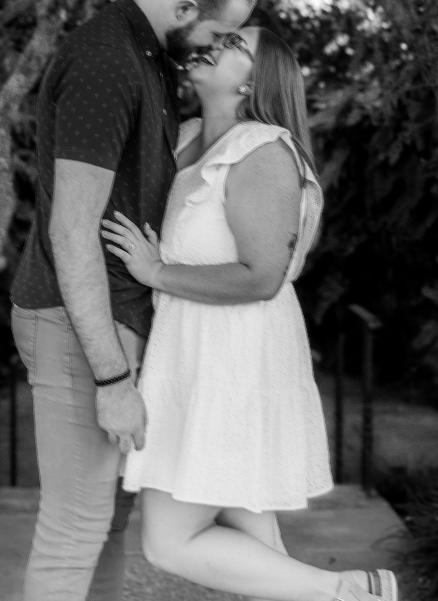 A couple in casual attire shares an intimate black and white kiss outdoors in a garden setting.