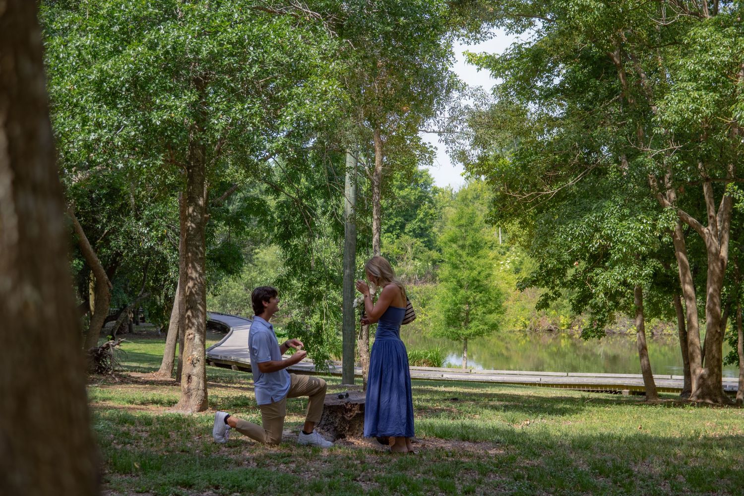 Man proposing on one knee to woman in blue dress among lush green trees near a river.