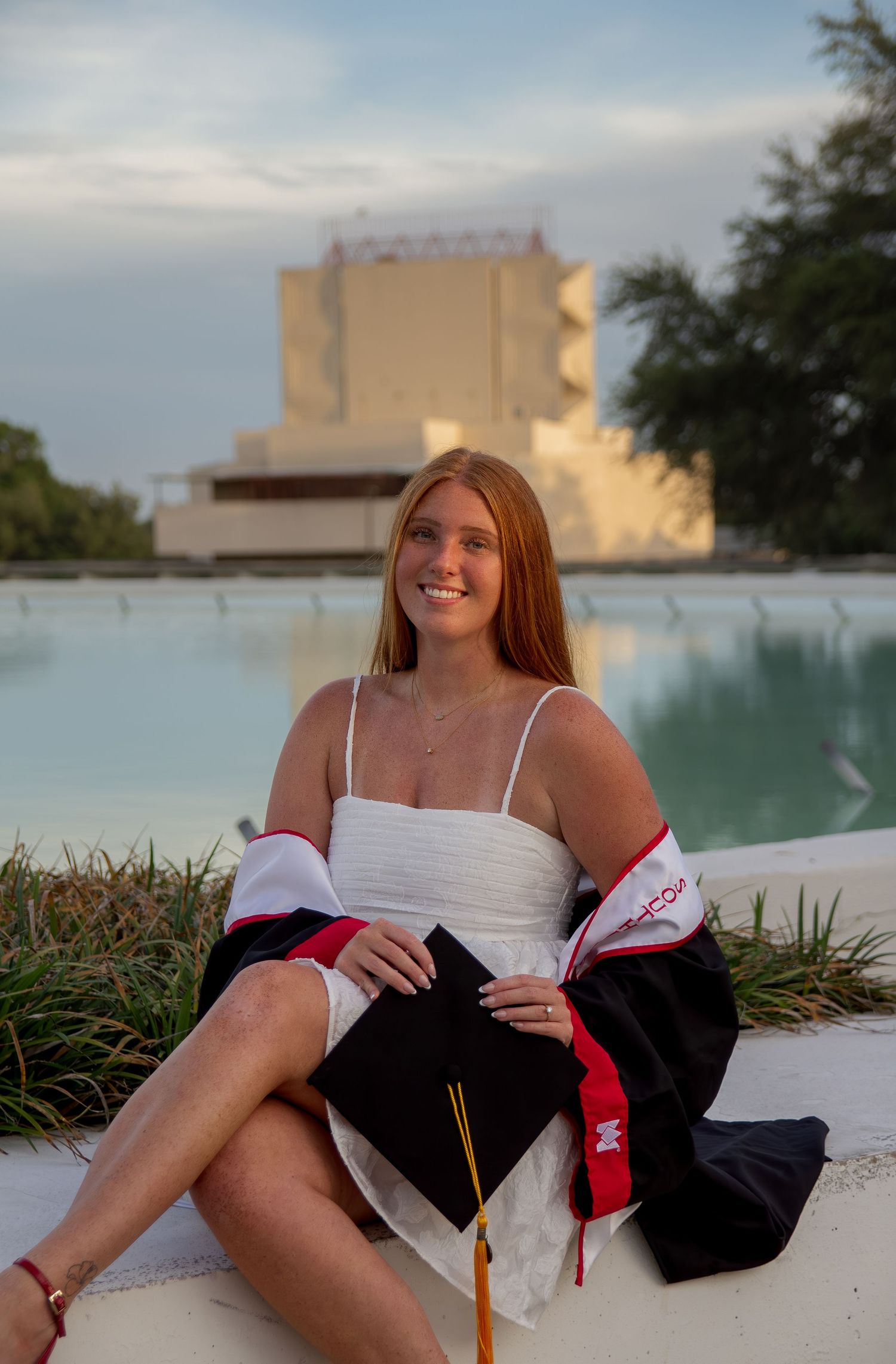 Smiling graduate in white dress holding black cap and gown by a pool at sunset.