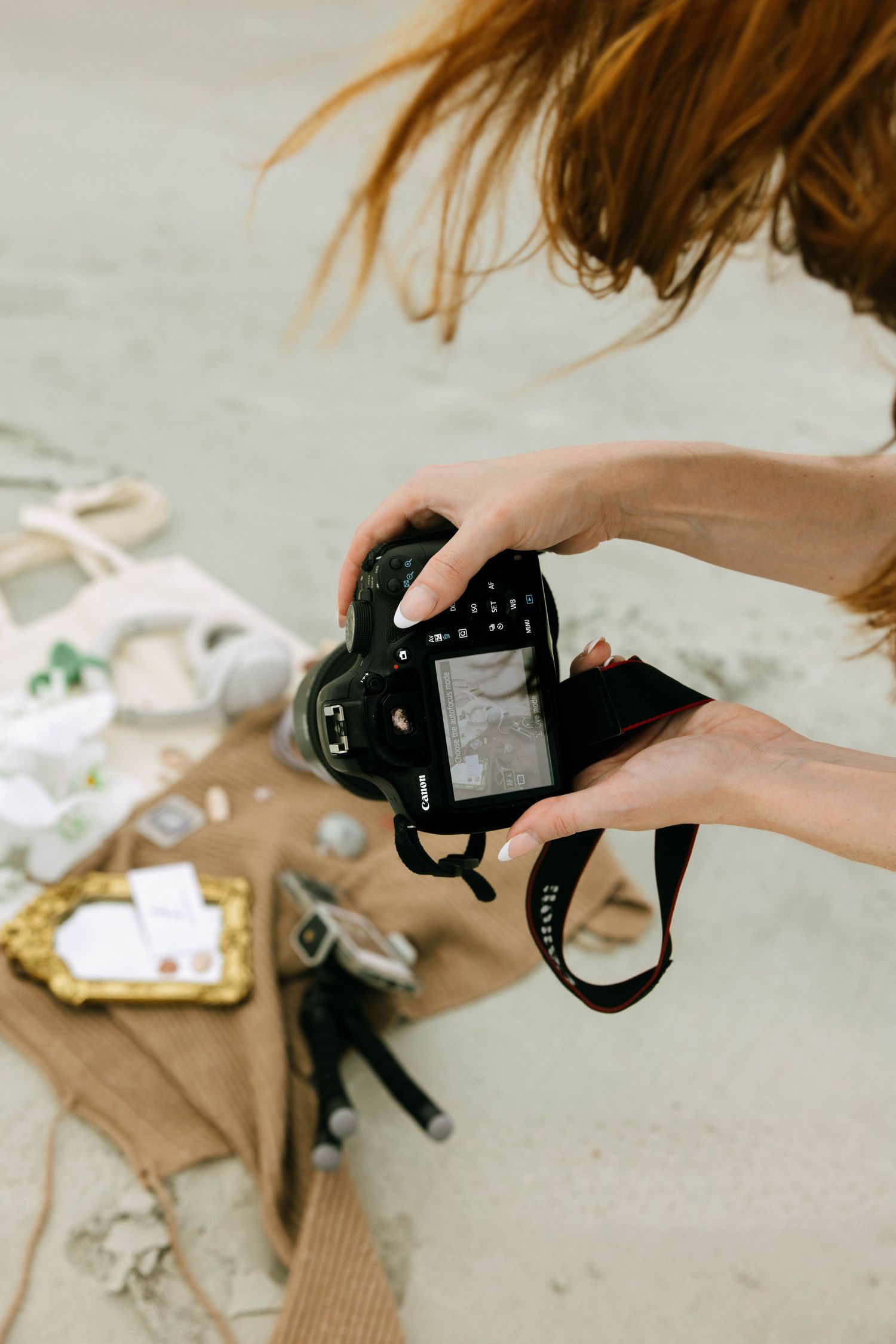 A redhead woman holds a Canon DSLR camera reviewing photos at a styled beach flat lay shoot.