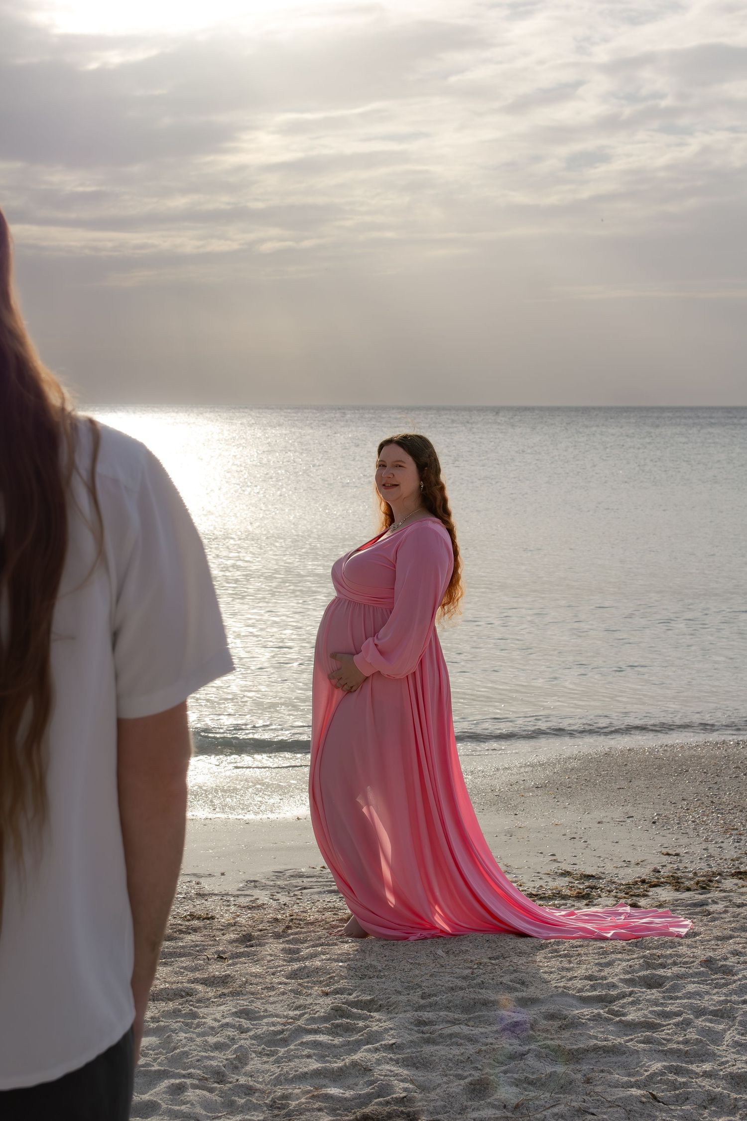 Pregnant woman in flowing pink gown poses on sandy beach at sunset near calm ocean waters.