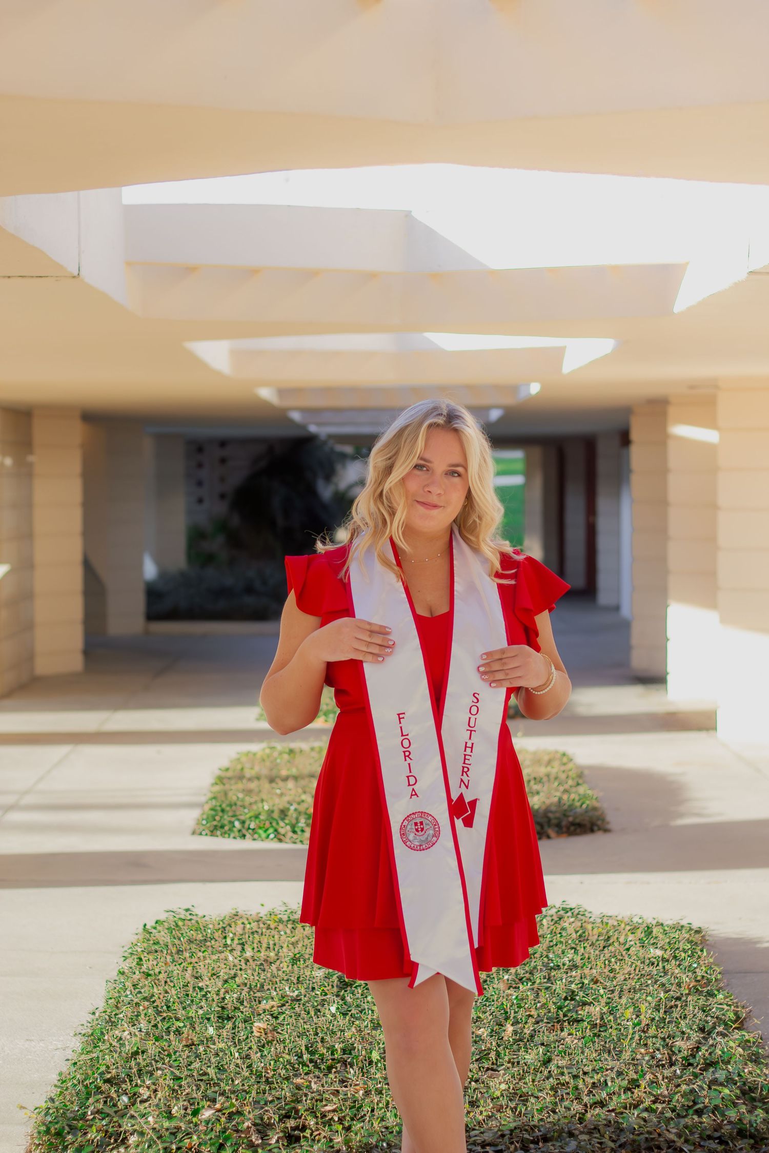 Graduate in red and white academic regalia poses outdoors near landscaping at a university campus.