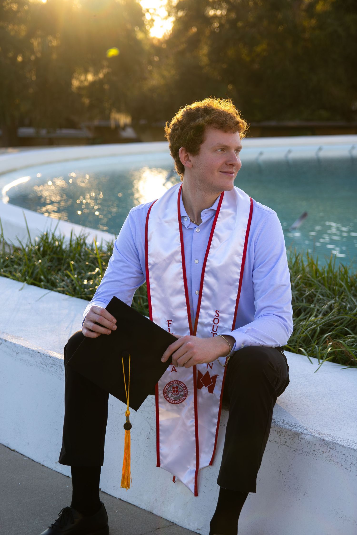 Someone in graduation attire sits on a white ledge at sunset by a pool while wearing red and white regalia.