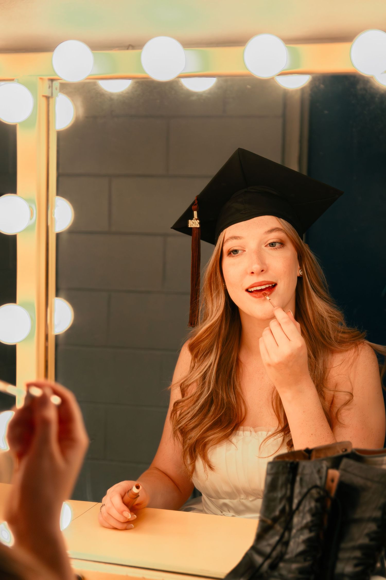 Graduate in cap and gown applies makeup in front of illuminated vanity mirror with round bulbs.