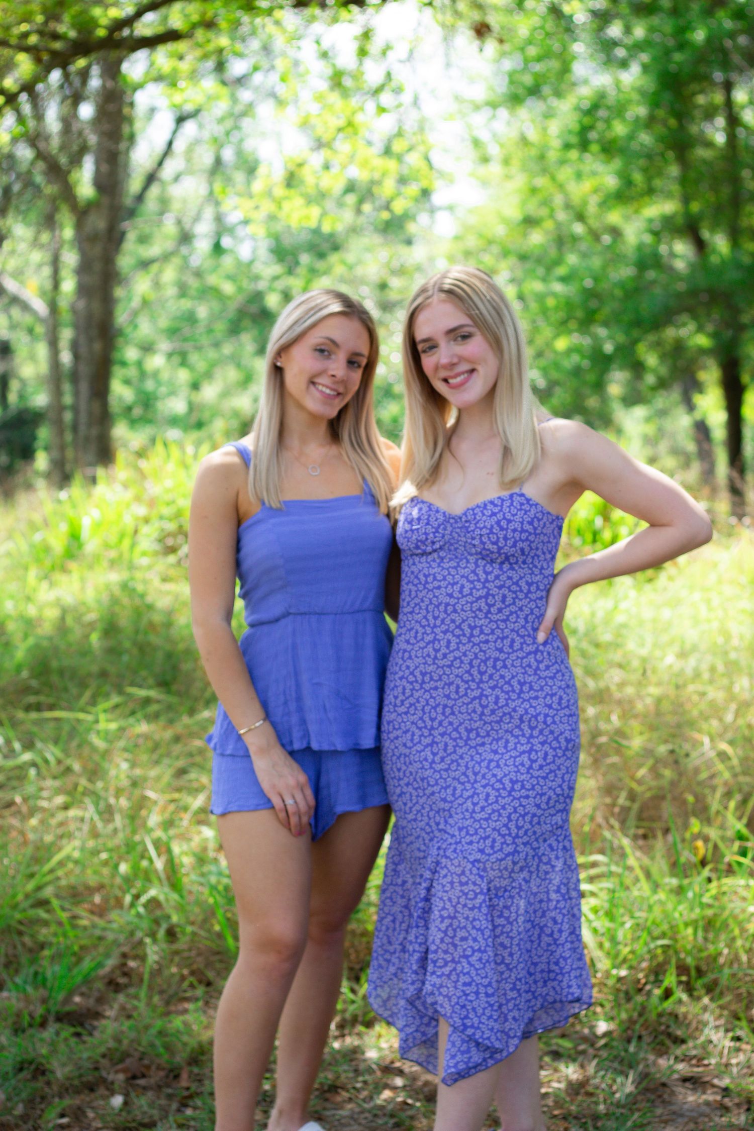 Two friends in summer outfits pose together in a sunny wooded area with green foliage in the background.