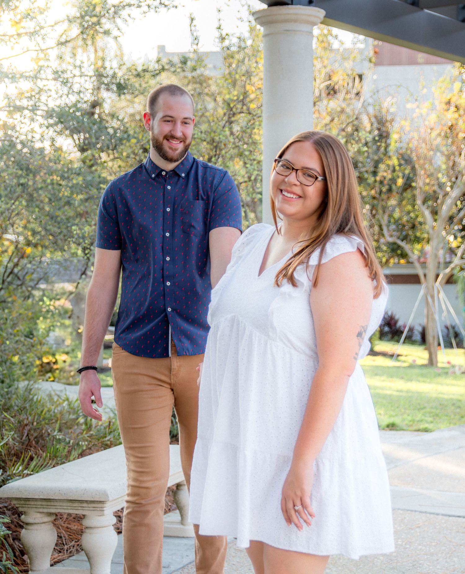 Casual couple stands together on sunny porch with white columns and garden setting.