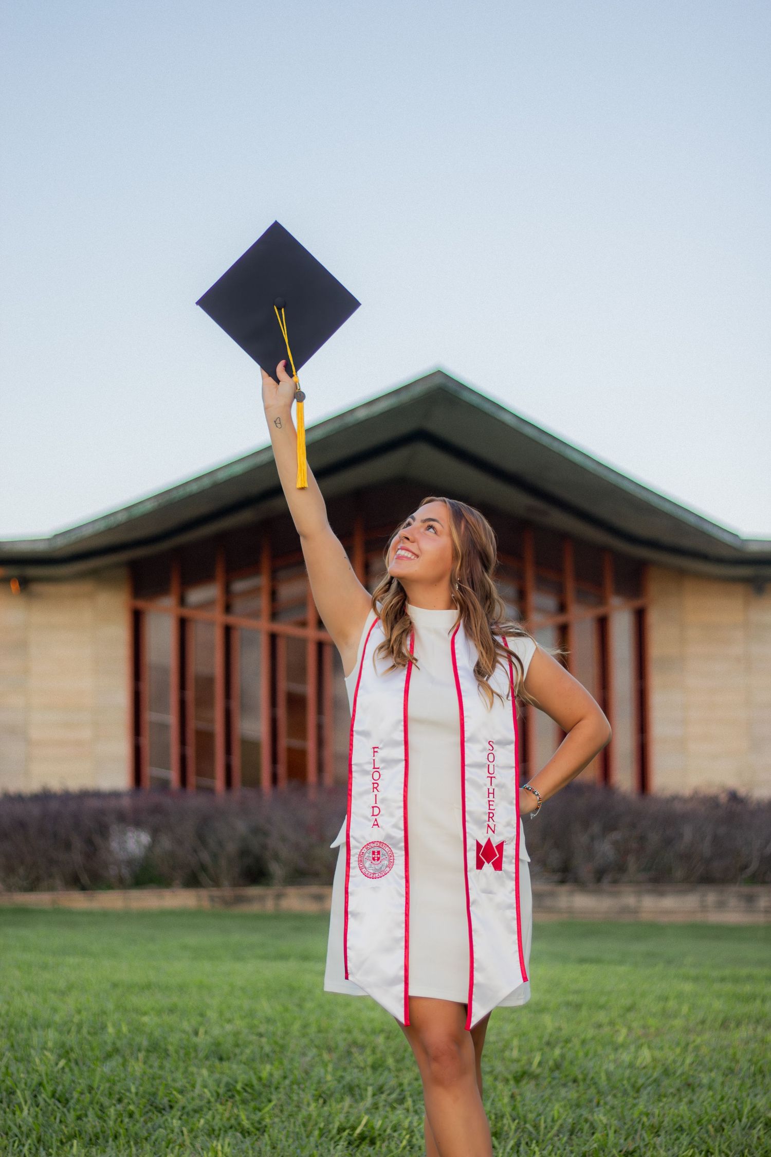 Graduate in white dress and stole throwing graduation cap in air by campus building.