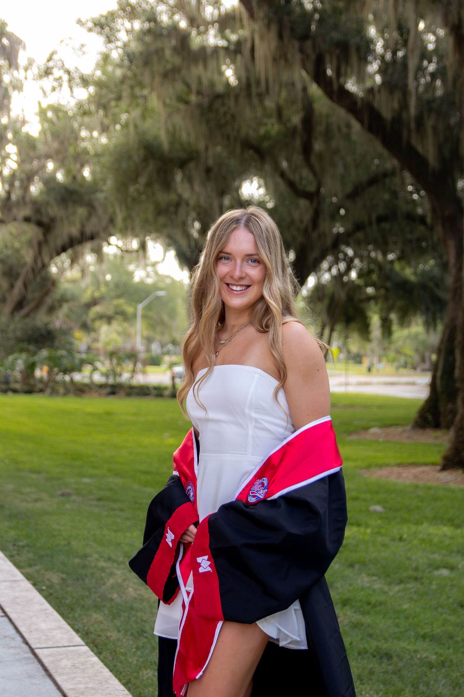 Smiling blonde woman in white dress holding graduation gown outdoors under moss-covered trees on a green campus.