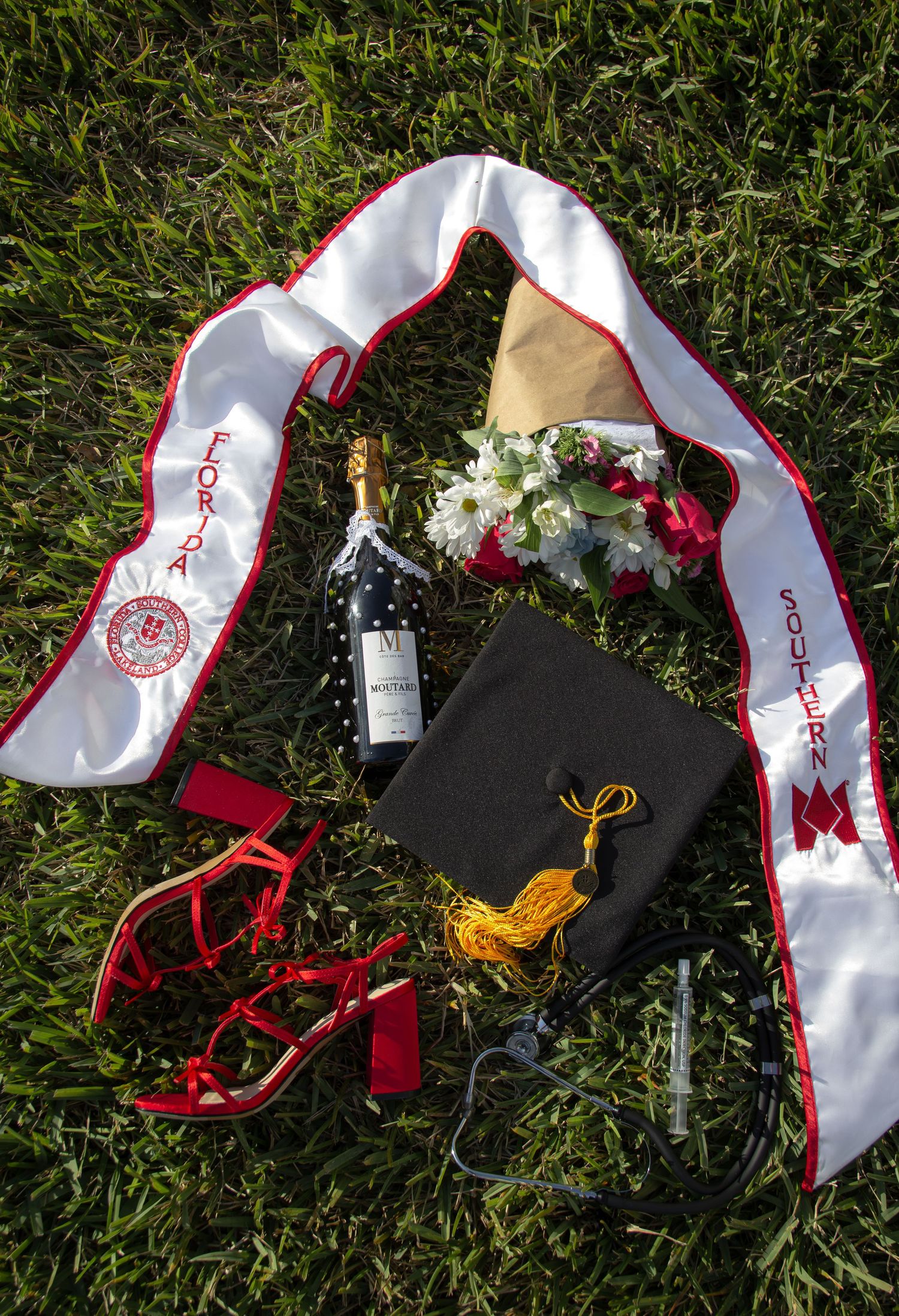Graduation flat lay with mortarboard, sash reading Fort Southern, wine bottle, flowers, and red heels on green grass.