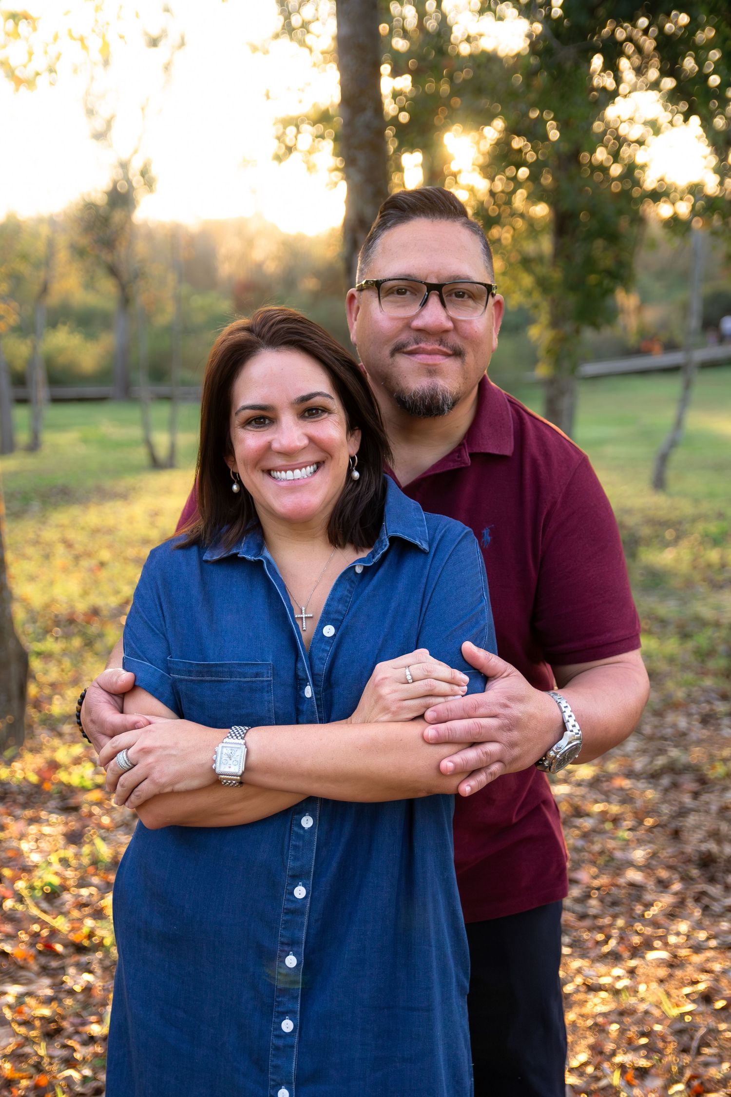 A couple embraces outdoors in autumn with golden sunlight filtering through trees and fallen leaves on the ground.