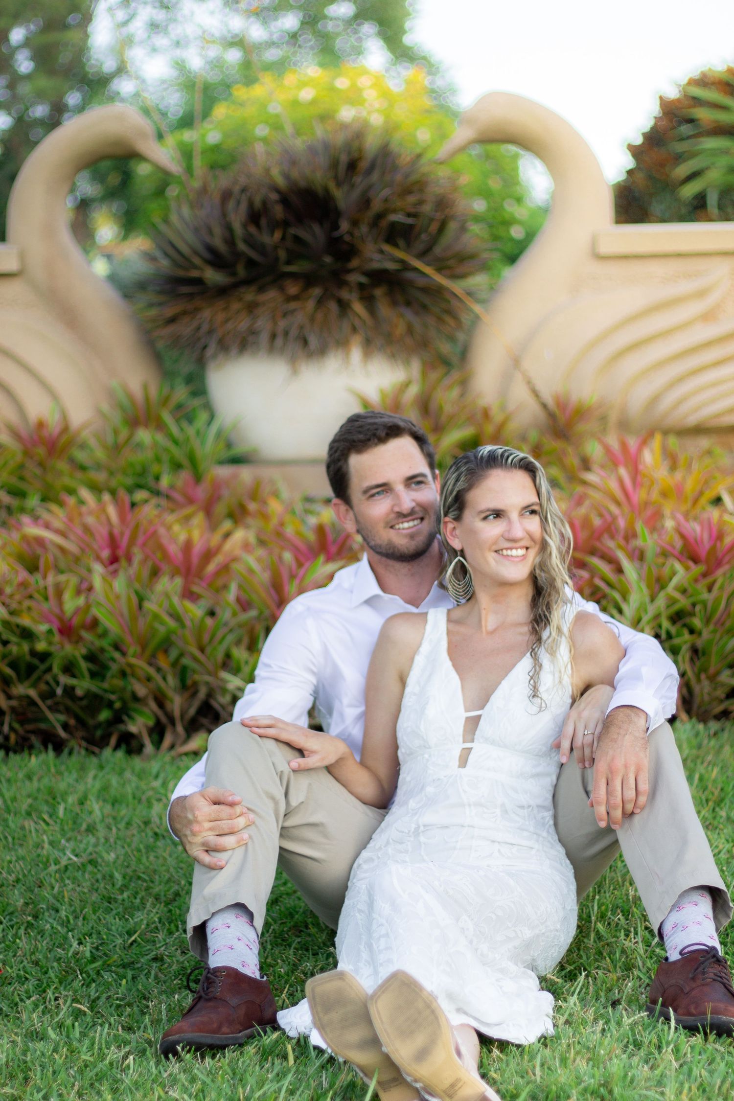Couple relaxes on grass near tropical landscaping in casual wedding attire.