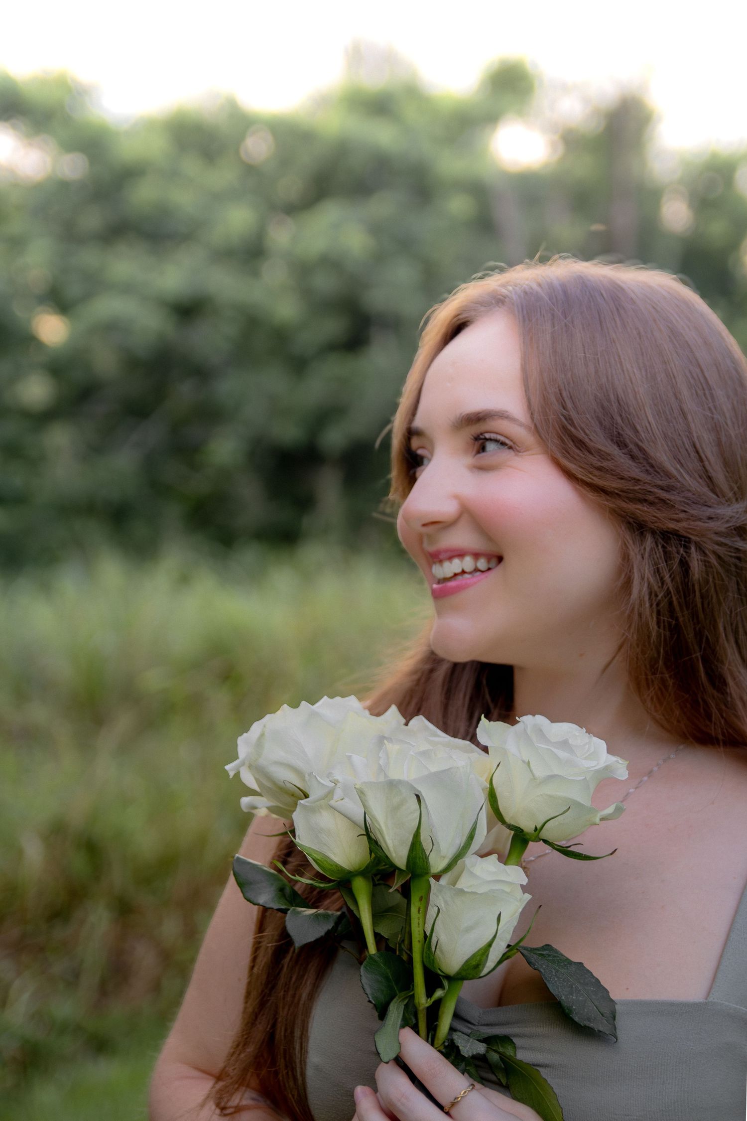 Woman in grey dress smiles while holding white roses outdoors in a natural garden setting during golden hour.
