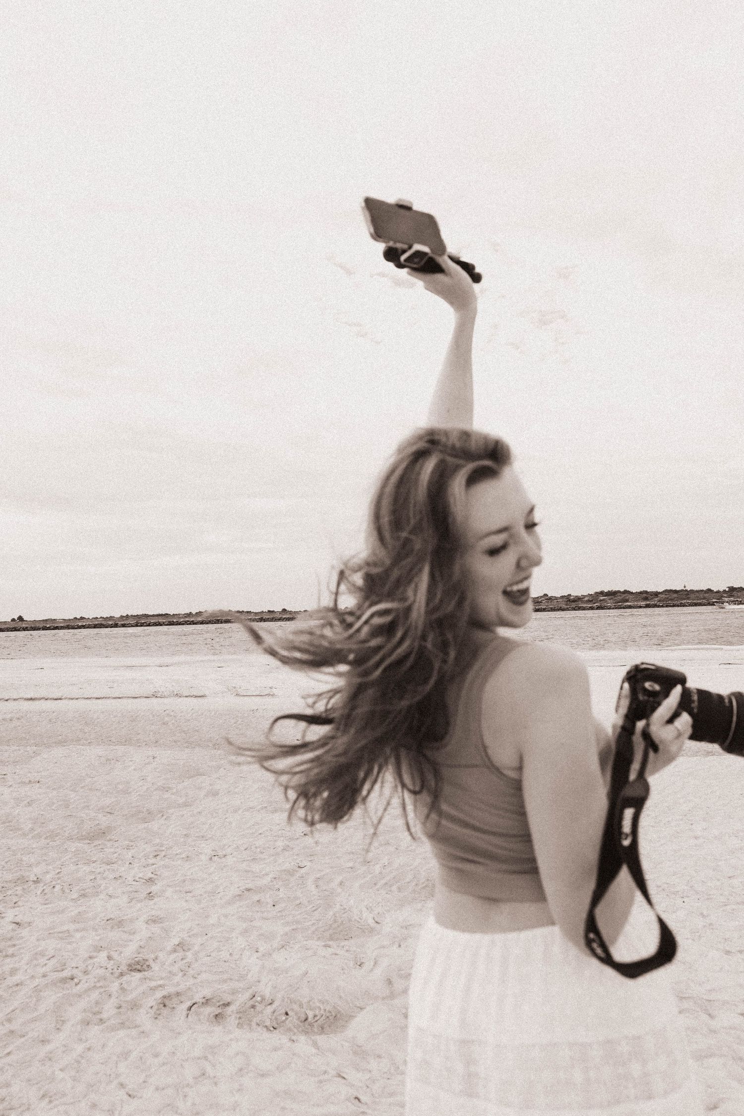 Young woman laughing on beach holding camera and phone above head, hair blowing in wind, black and white photo.