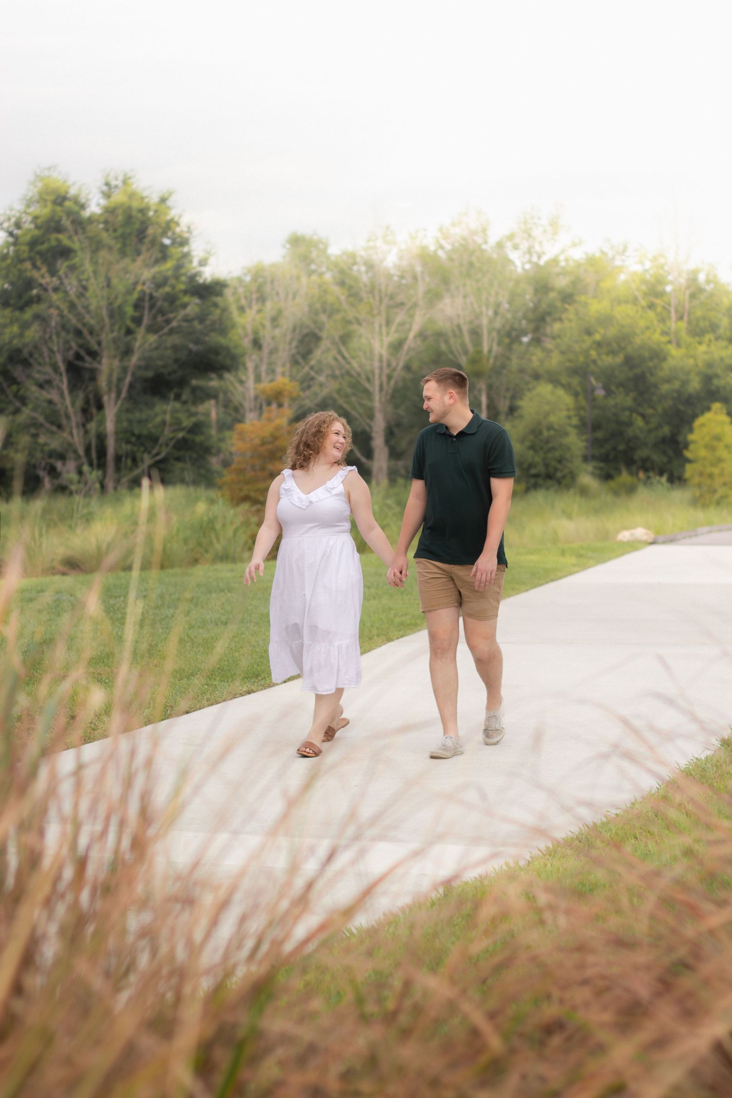 Couple walking hand in hand along concrete path with tall grass.