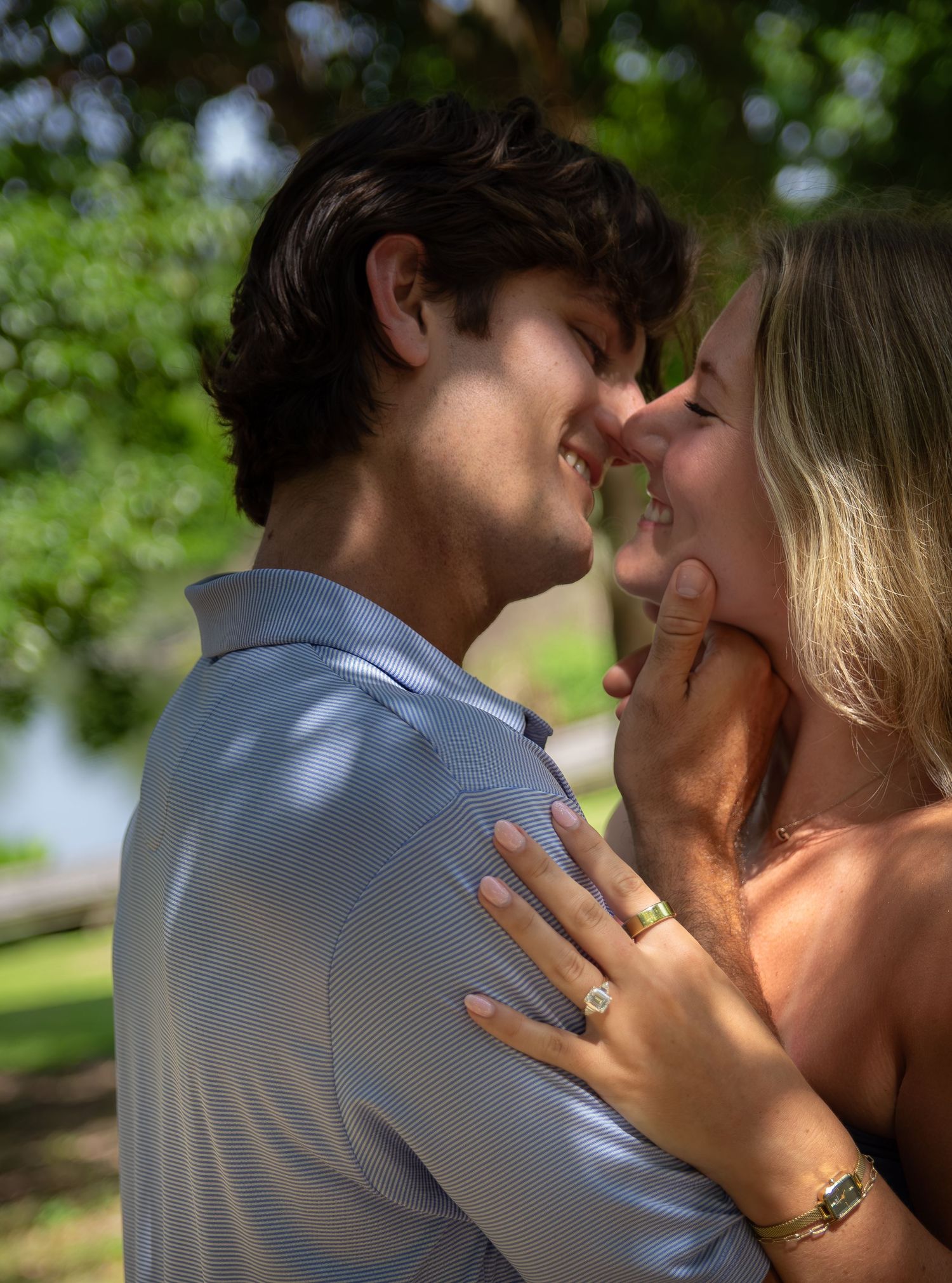 Couple sharing a romantic moment outdoors, woman displaying engagement ring, surrounded by lush green trees.