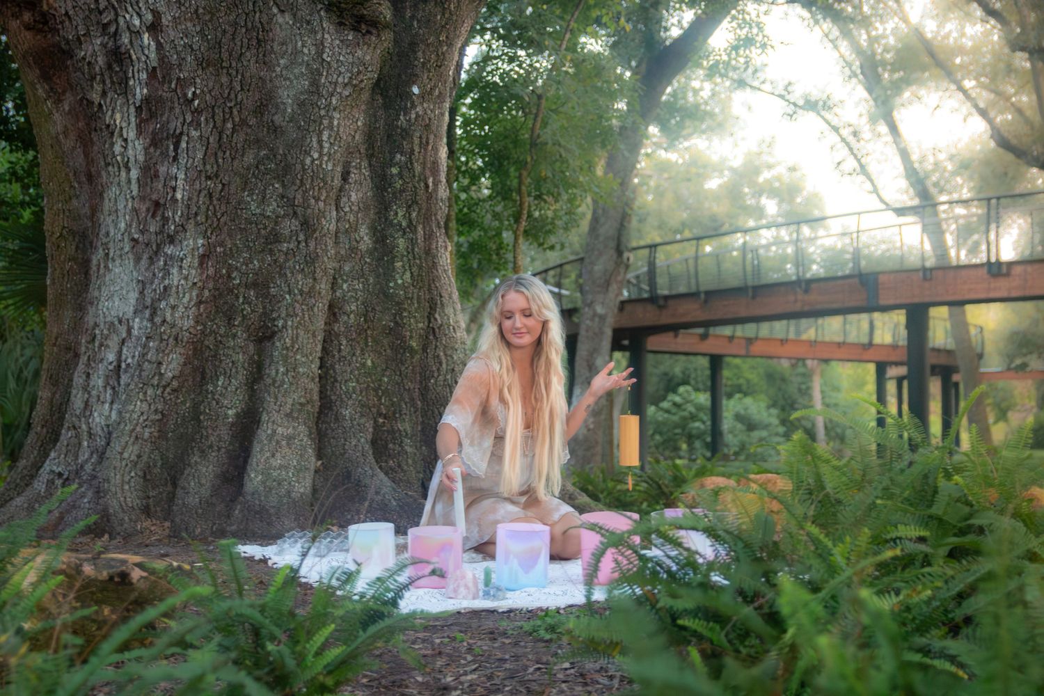 A dreamy outdoor picnic scene with a pink blanket set beneath massive trees near a wooden footbridge in dappled sunlight.