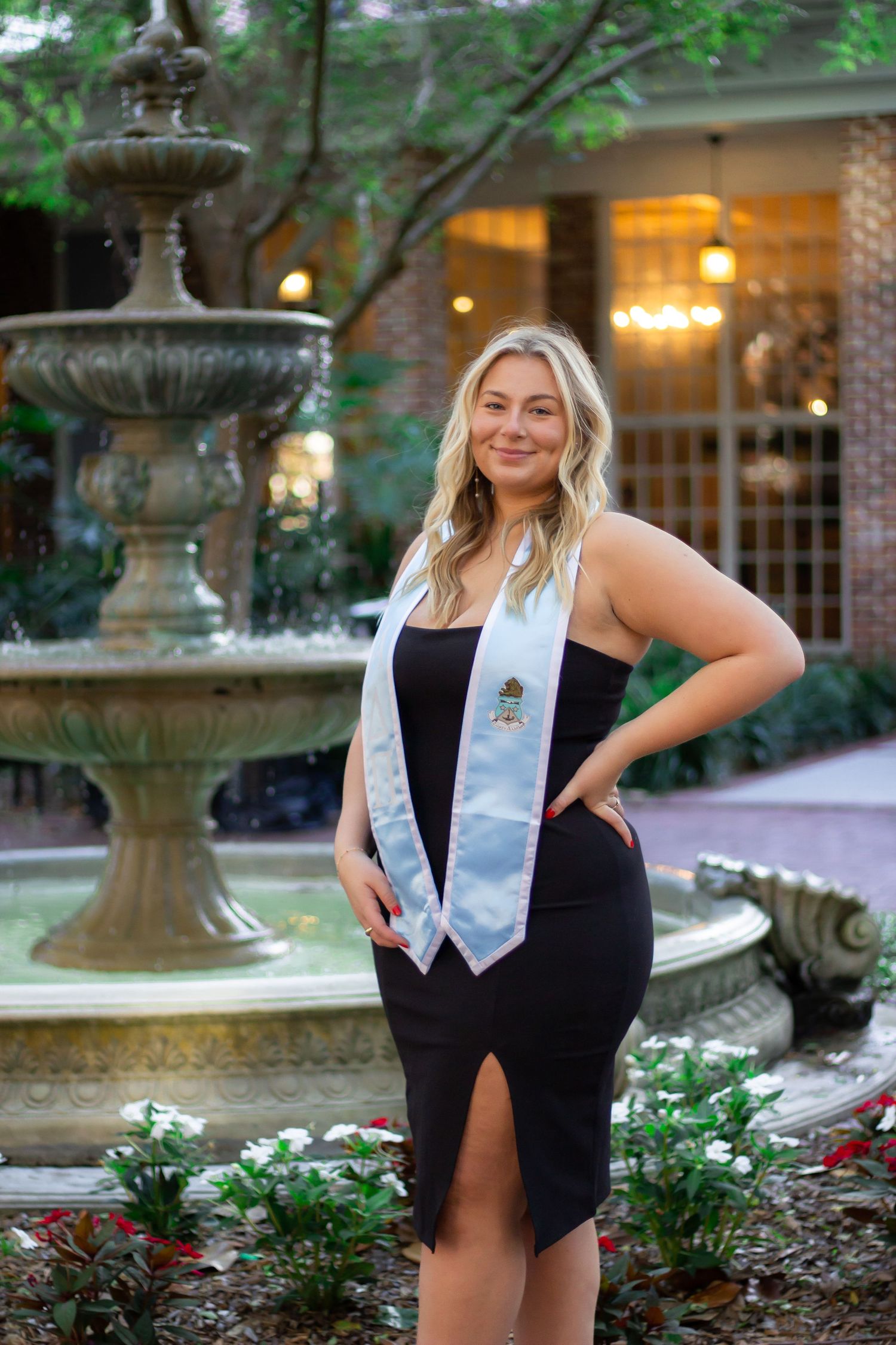 Graduate poses in black dress and white sash near ornate garden fountain during outdoor photo session.