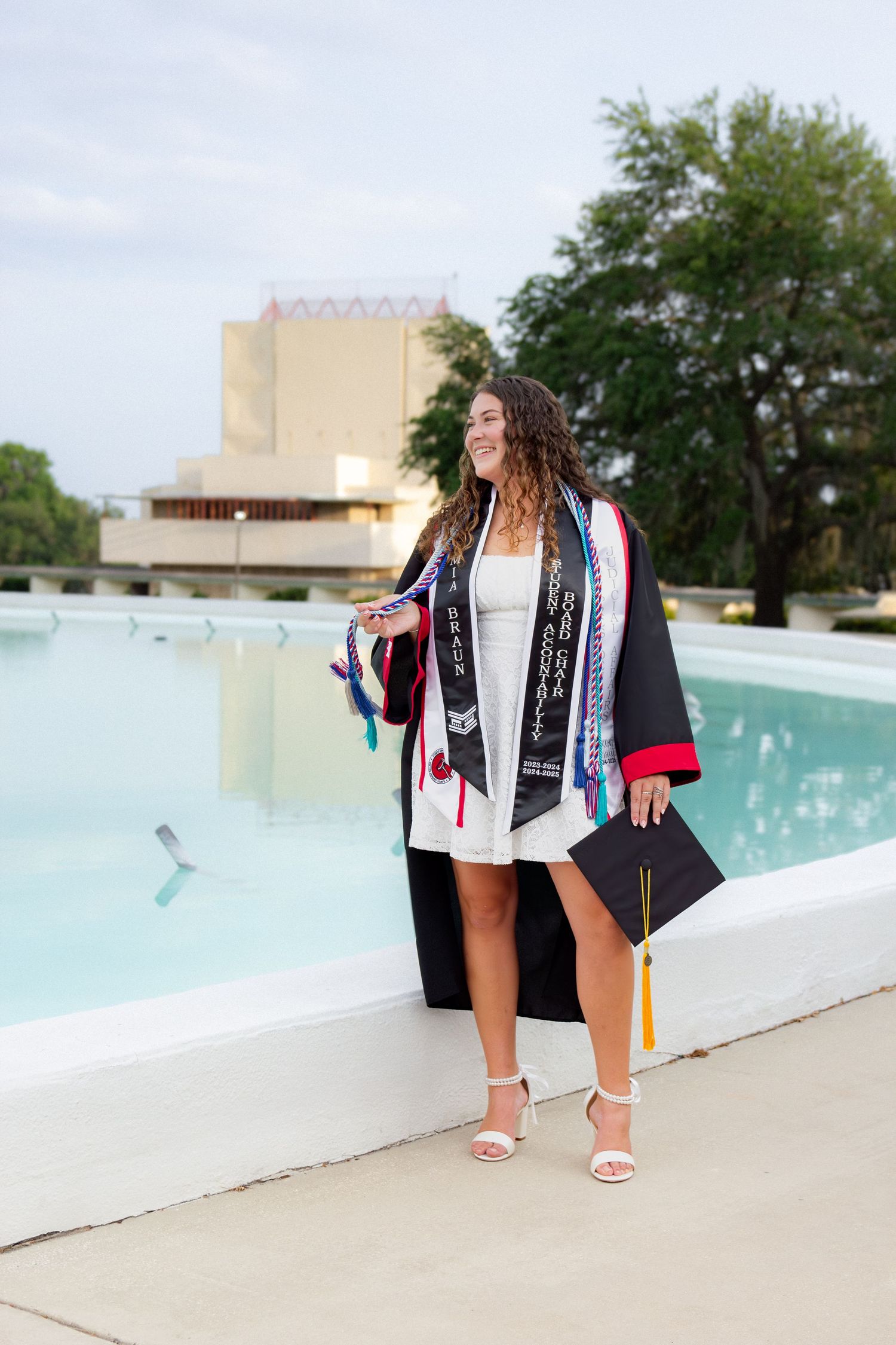 Graduate posing in cap and gown by a poolside on graduation day.