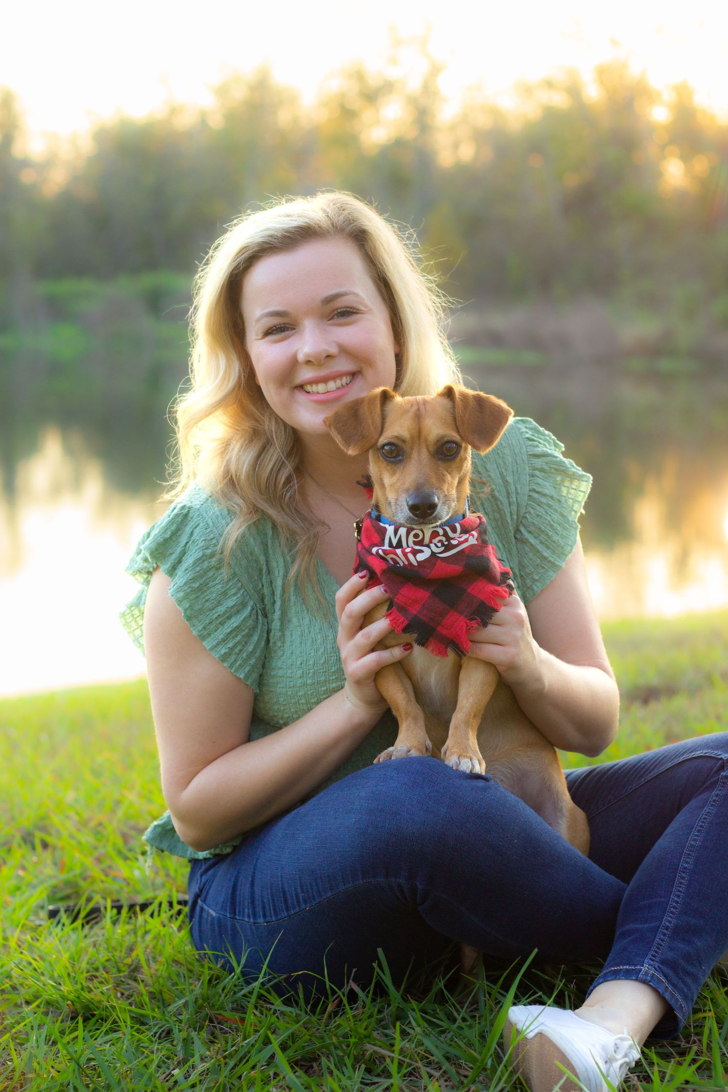Small dog wearing red bandana being photographed on grass near waterfront at sunset.