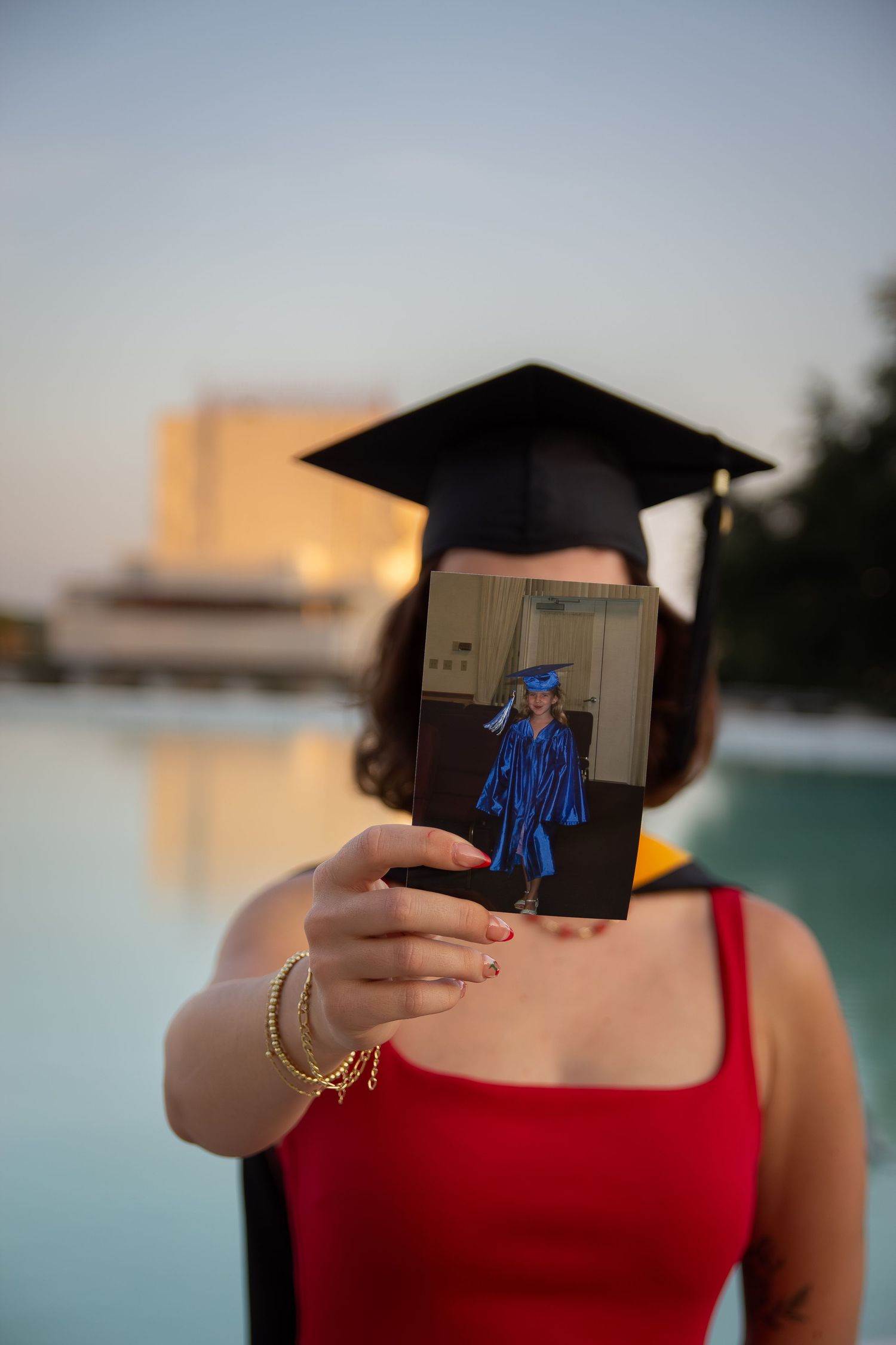 Graduate in red dress and mortarboard holds childhood graduation photo outdoors near pool at sunset.