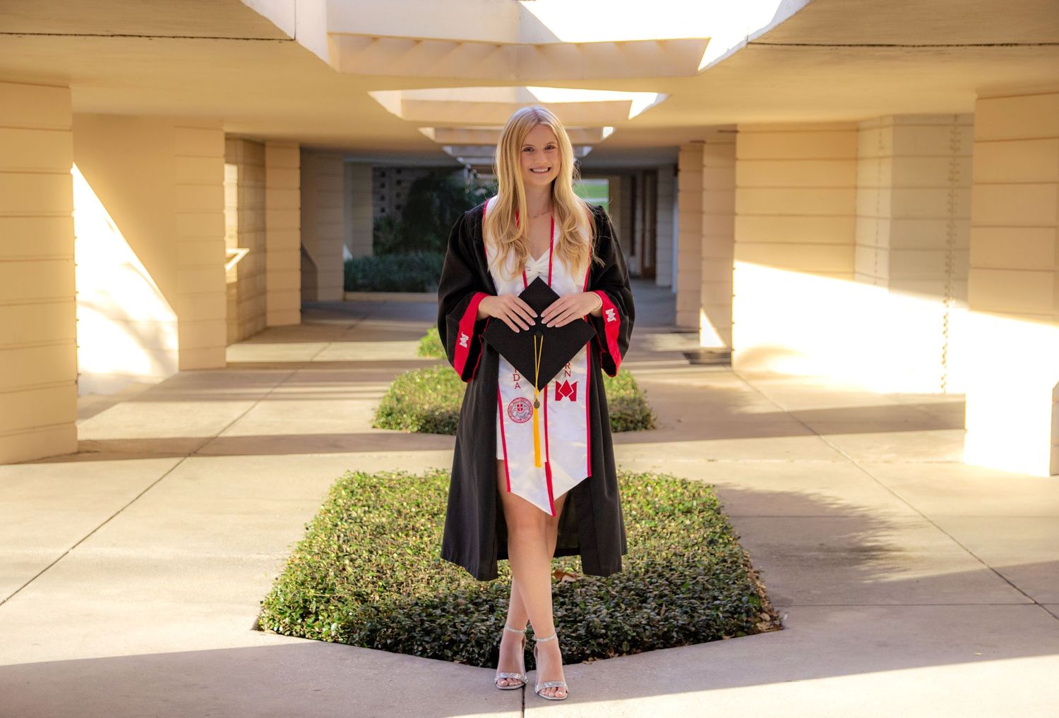 A person in a black dress stands in a series of architectural archways with beige columns and landscaping.