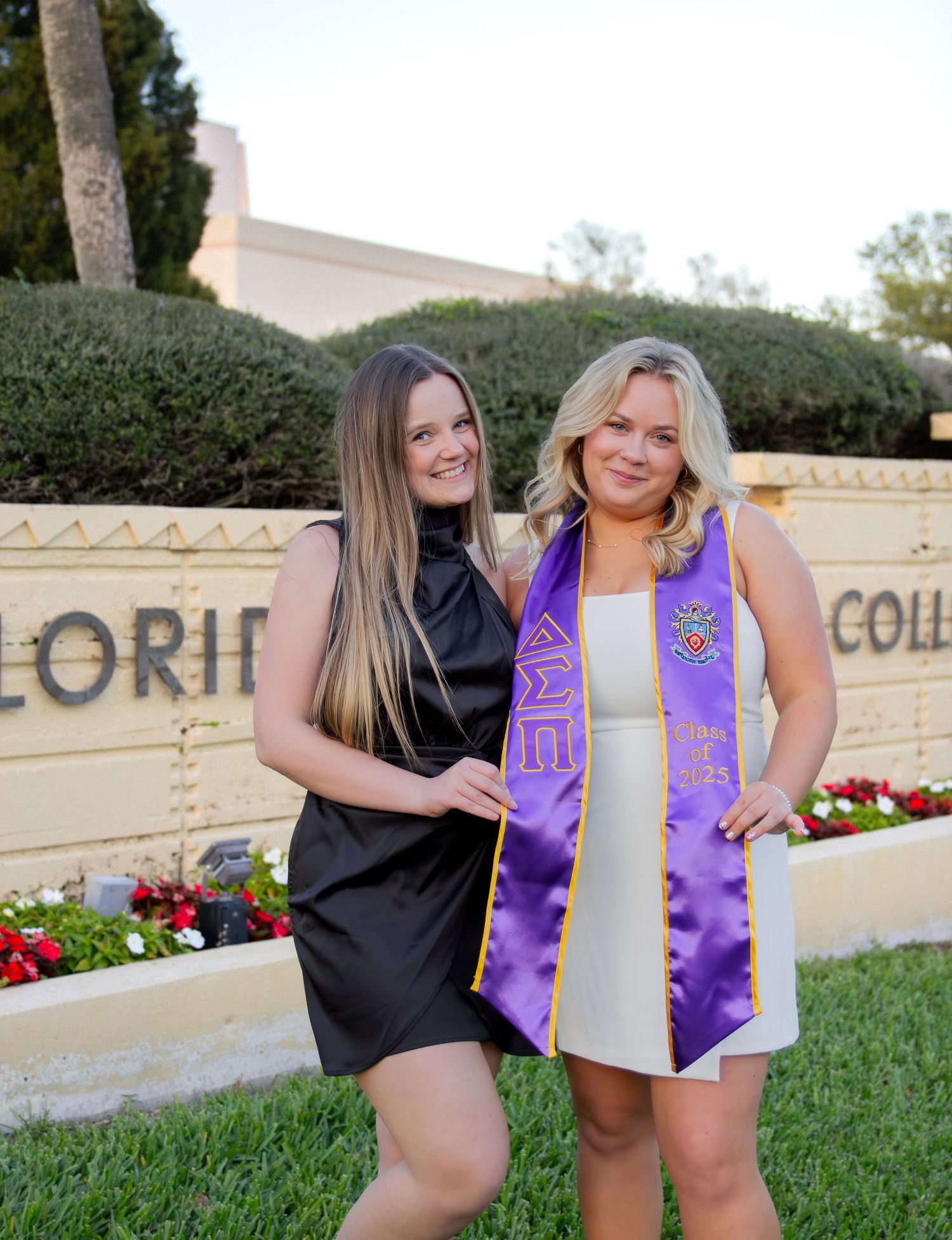 Two graduates in purple and white sashes pose together on a grassy lawn in front of a stone wall with flowers.