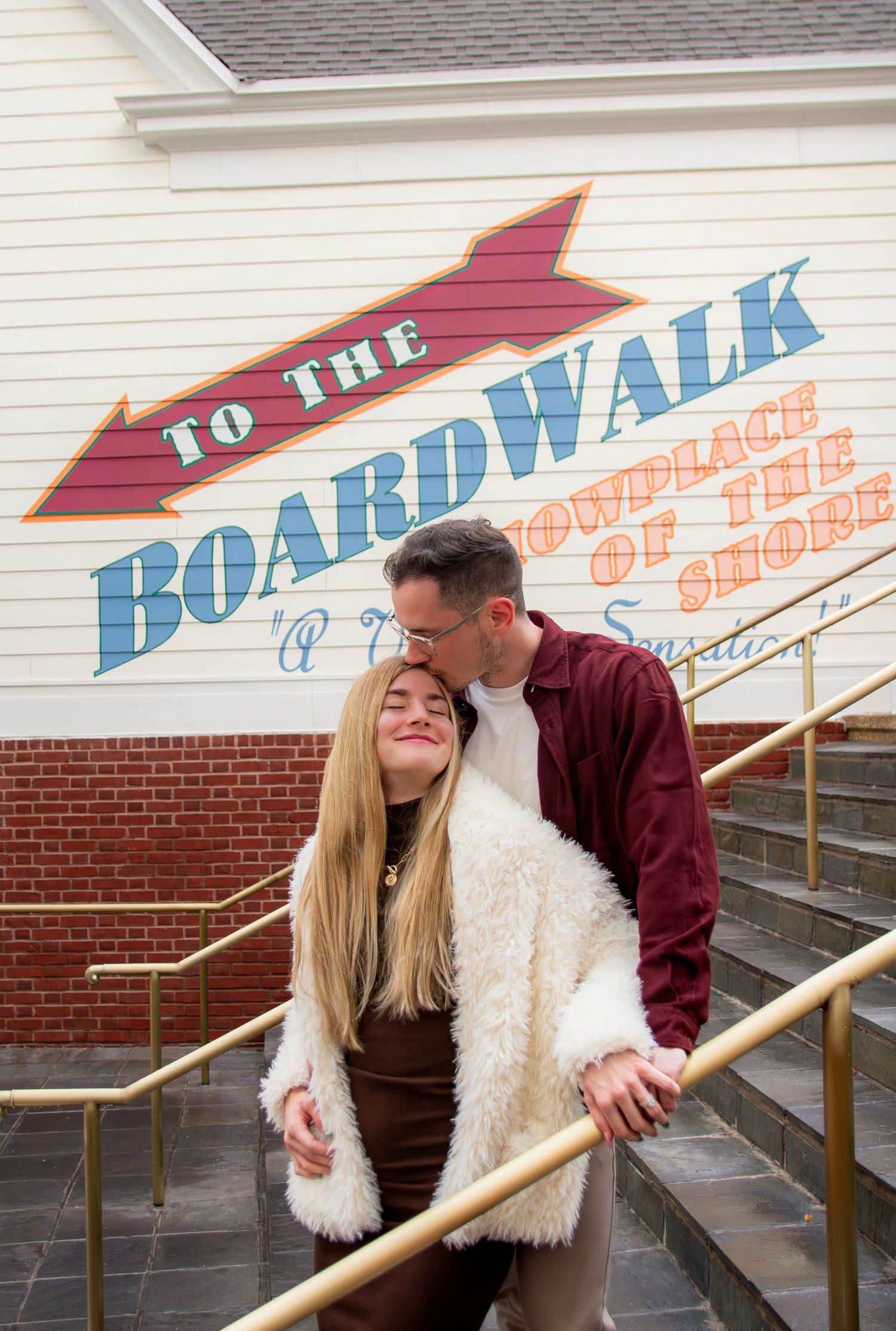 A couple shares a playful moment on stairs near a boardwalk sign, with one wearing a white fuzzy coat.