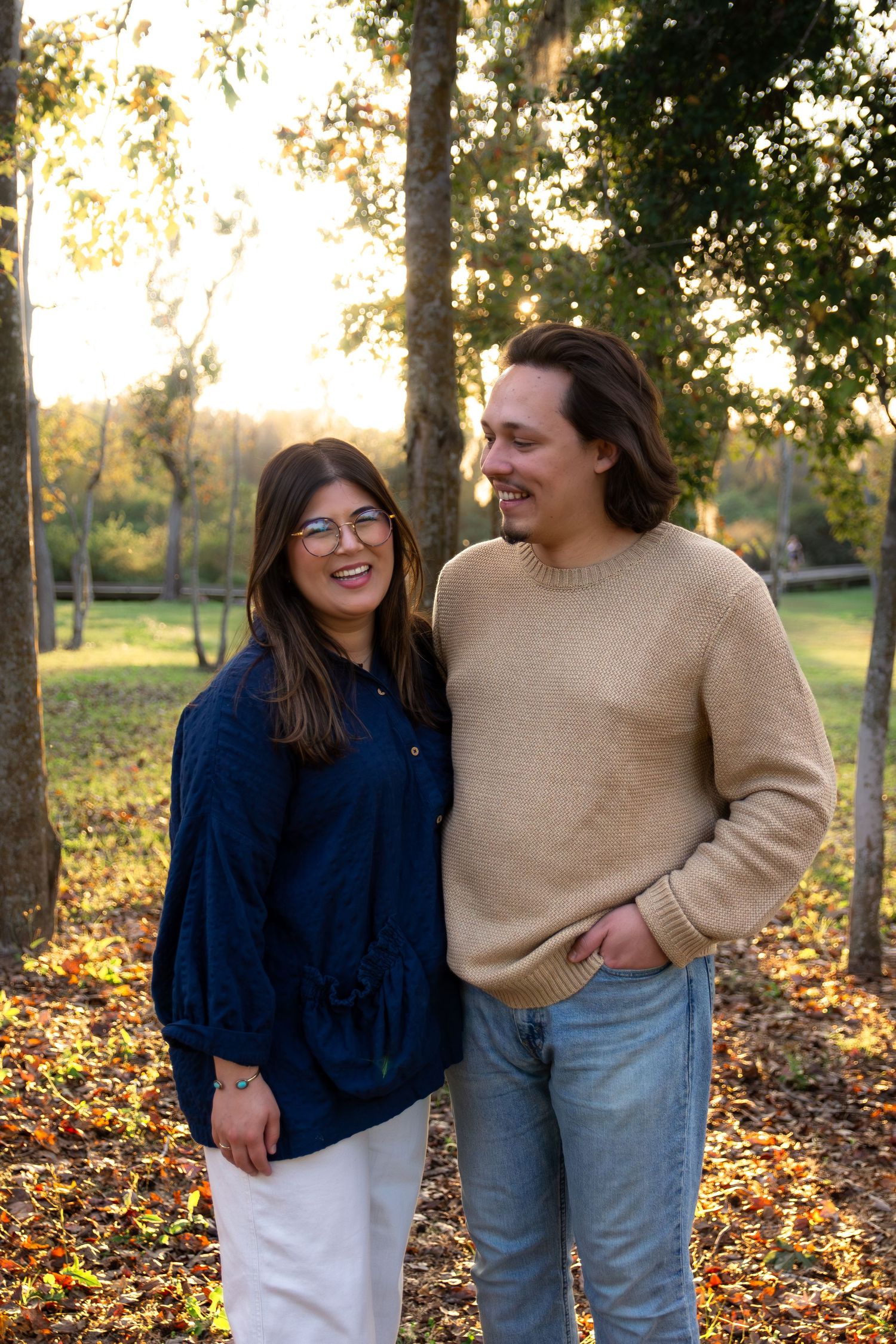 A couple laughs together while standing among trees in a park during golden hour with autumn leaves scattered below.