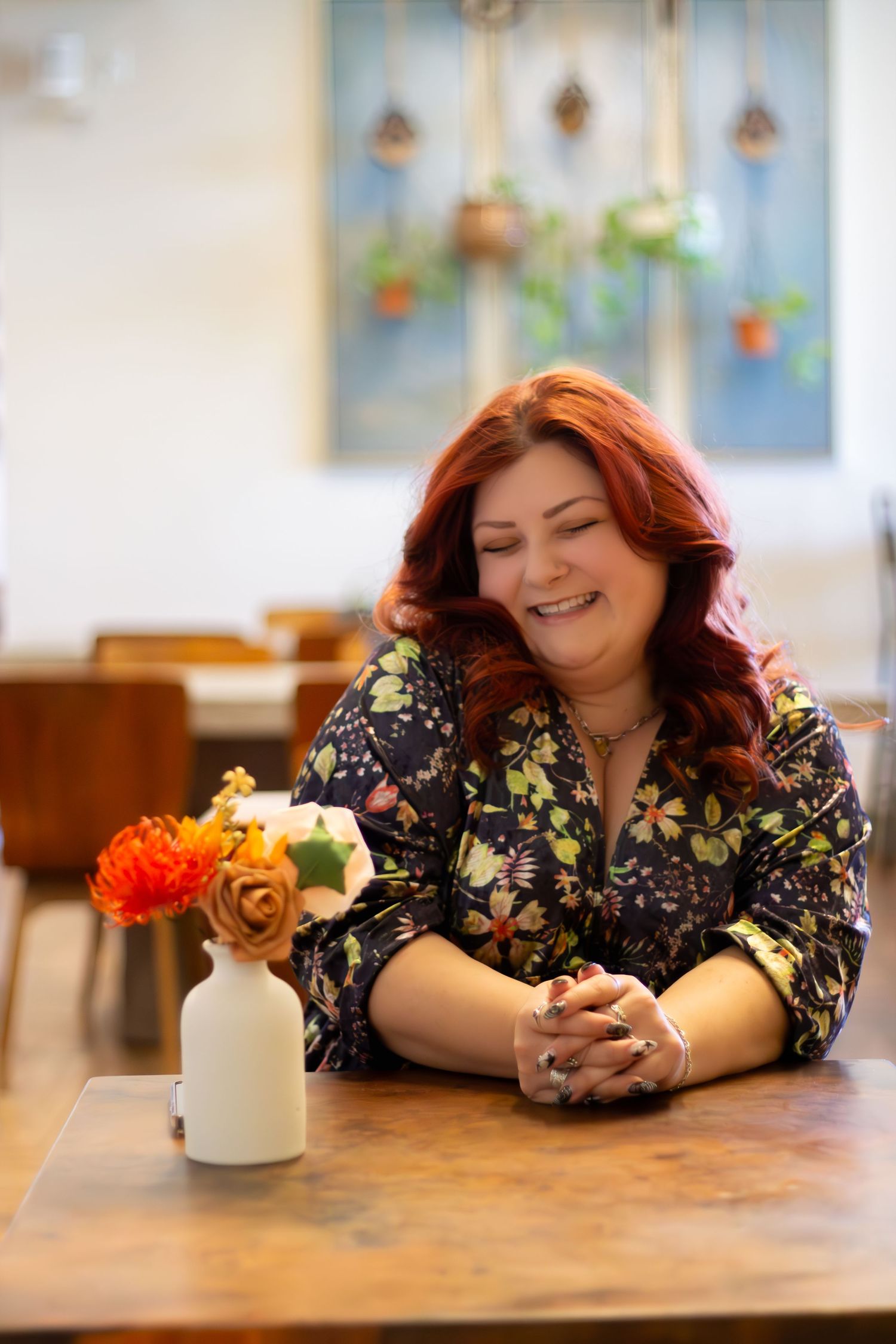 A person smiles while looking down at a phone at a wooden table with flowers in the background.