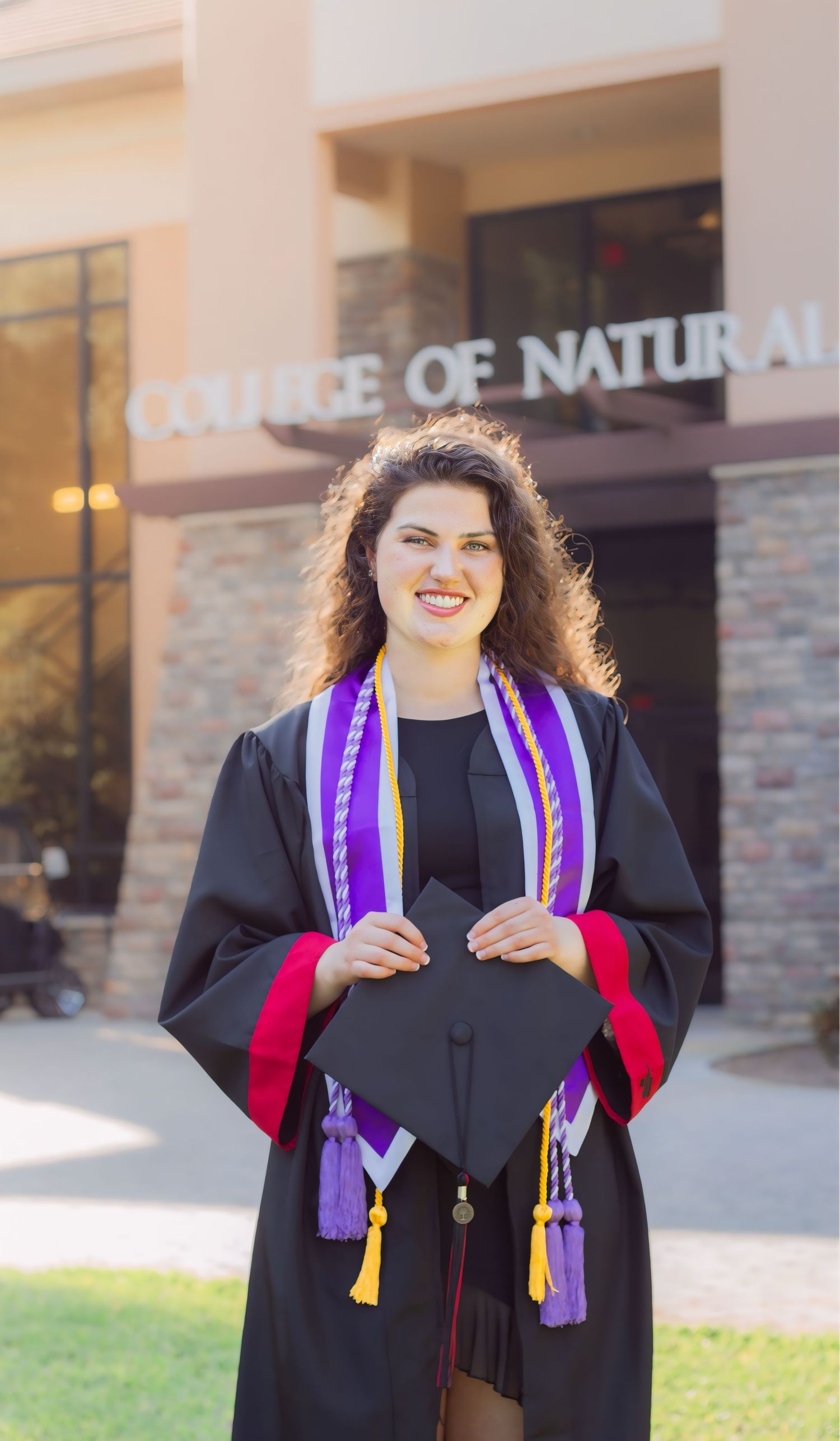 Graduate in black cap and gown with purple stole standing outside a building.