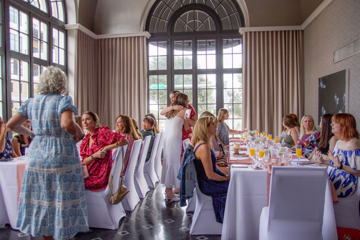 Guests mingle and socialize at an elegant event in a grand room with arched windows and white-clothed tables.
