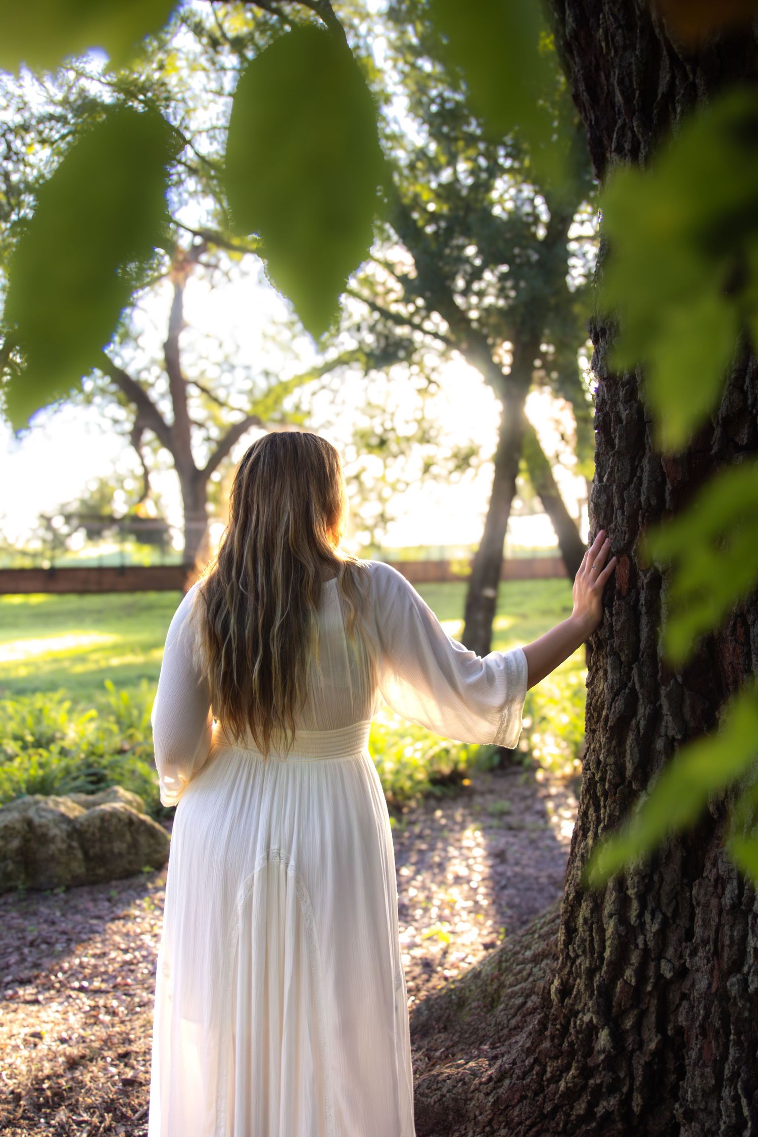 A person in a flowing white dress stands among trees during golden hour as sunlight filters through the leaves.