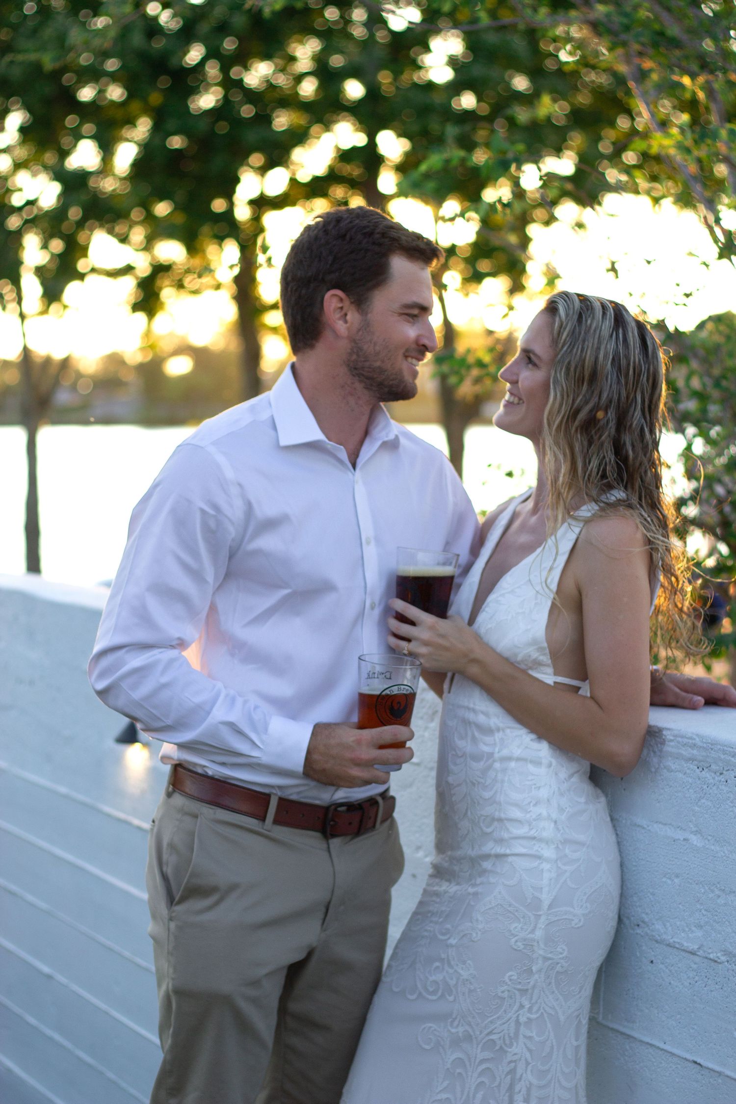 Couple shares drinks and kisses against white wall with garden backdrop.