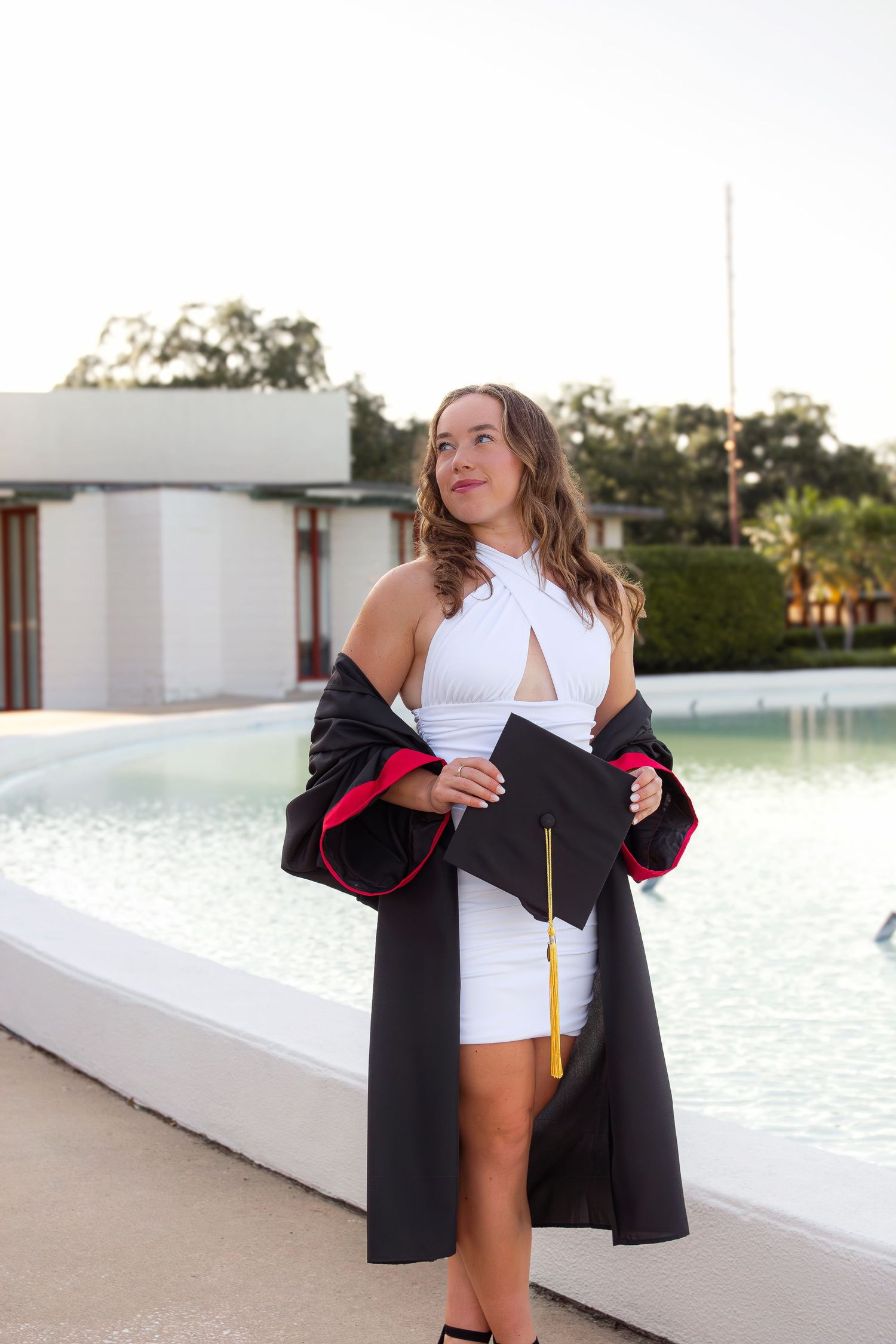 Graduate celebrating in cap and gown by waterfront during sunset photo session.
