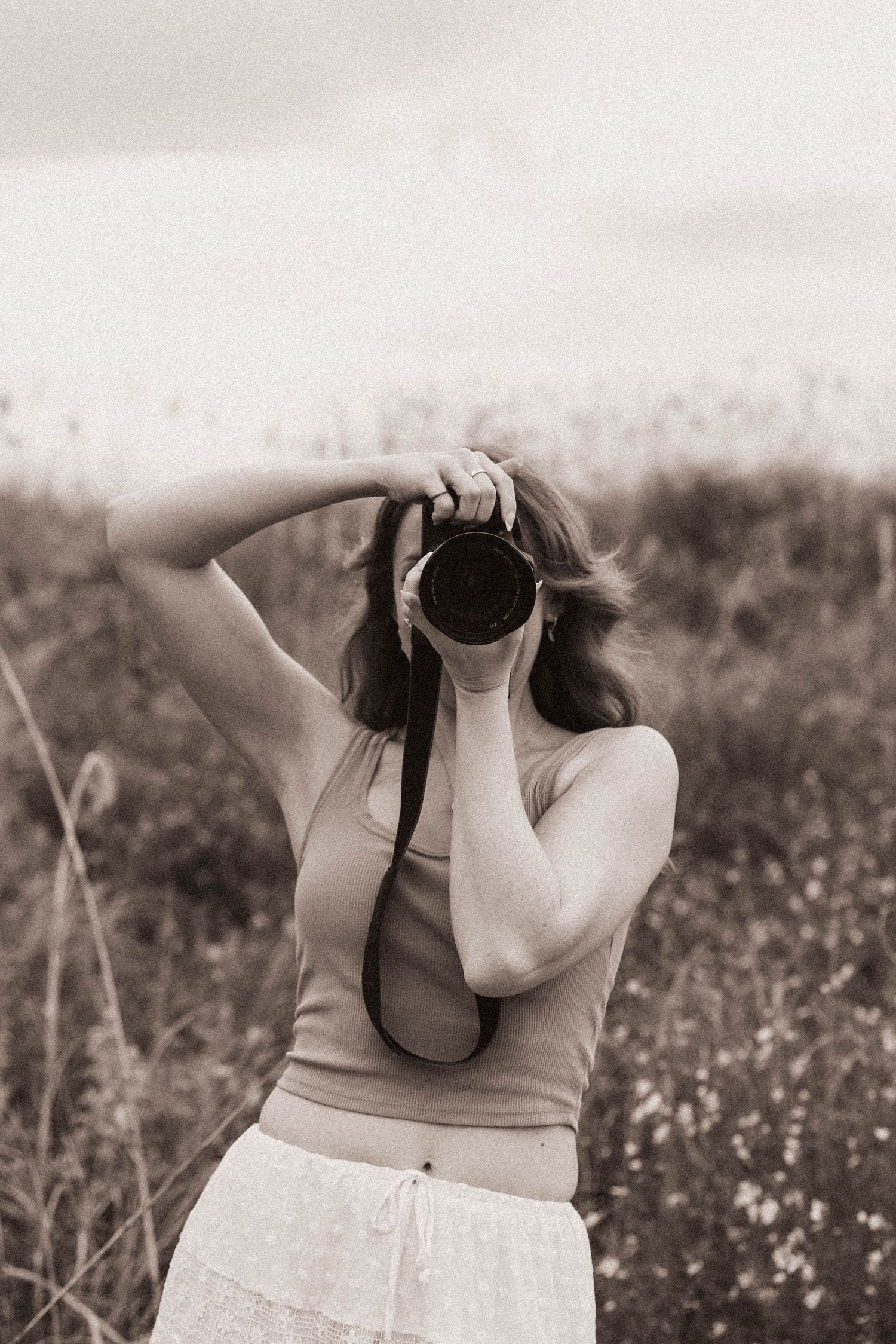 A woman holds a camera up to her face while standing in a field, captured in a vintage sepia-toned photograph.