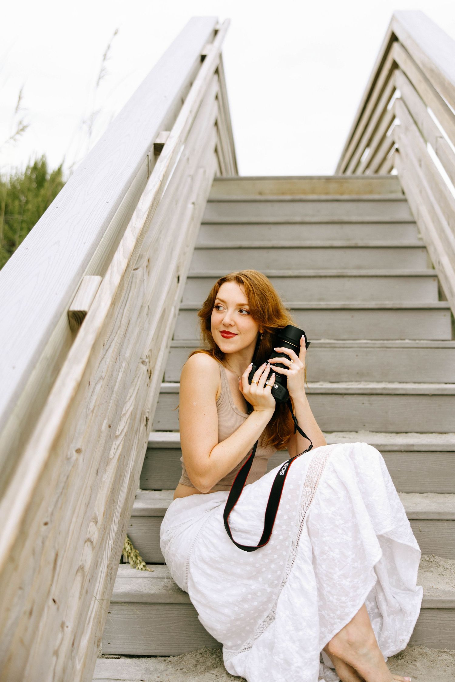 A young redhead woman in a white dress sits on wooden stairs holding a camera with a black strap.