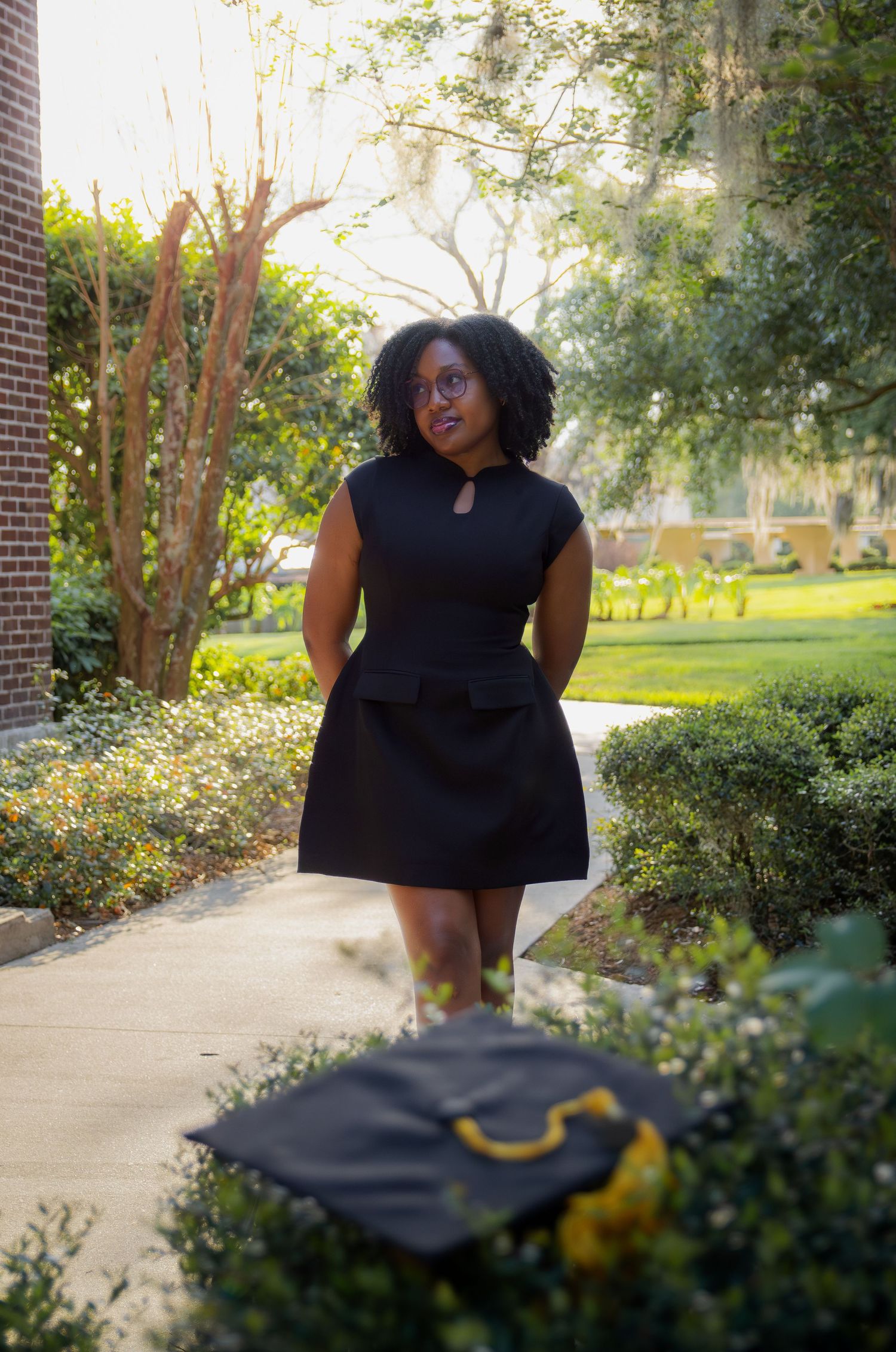 A young Black woman in a black peplum dress holds a graduation cap on a sunny campus walkway.